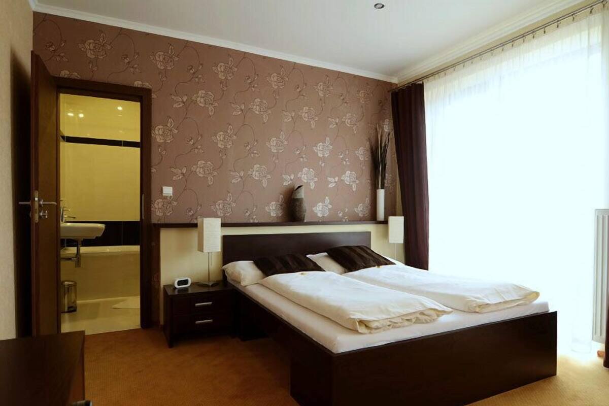 A hotel room with a large bed, a bedside table, a nightstand, a lamp, a wall-mounted shelf, and a bathroom visible through an open door.