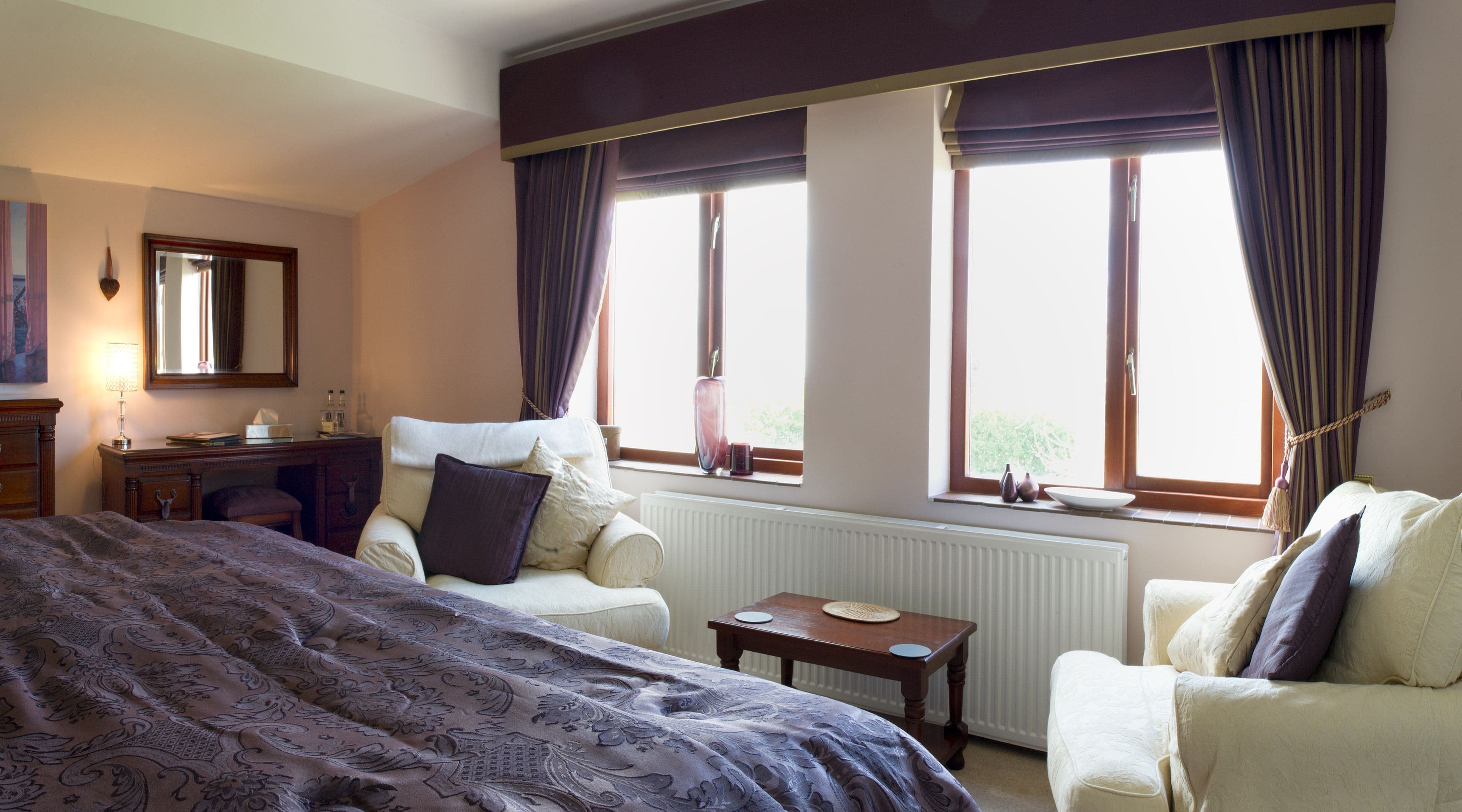 A hotel room with a large bed, two armchairs, a small table, a dresser, and a window with curtains.