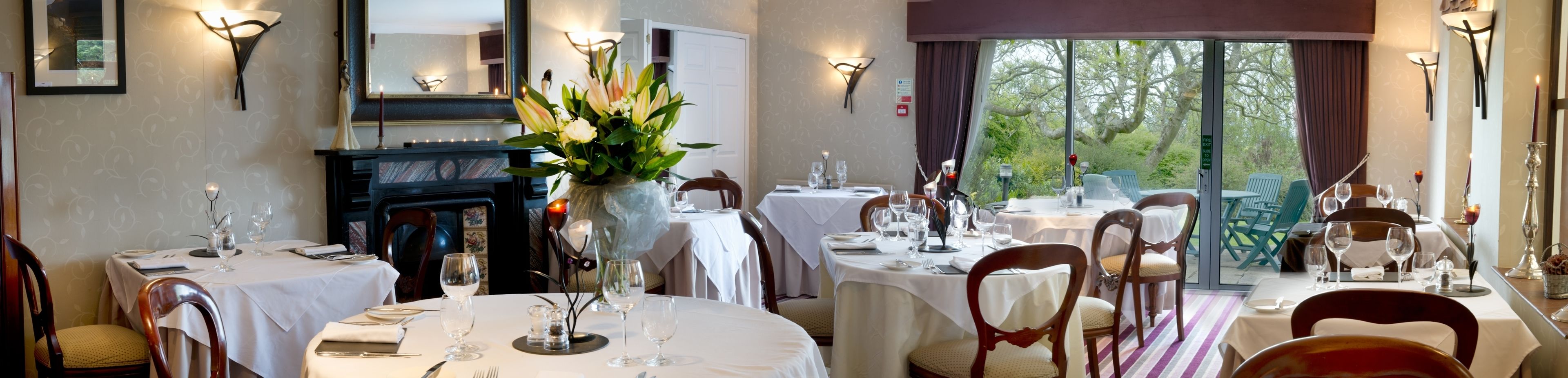A dining area with tables set for two, featuring white tablecloths, candles, and a vase of flowers.