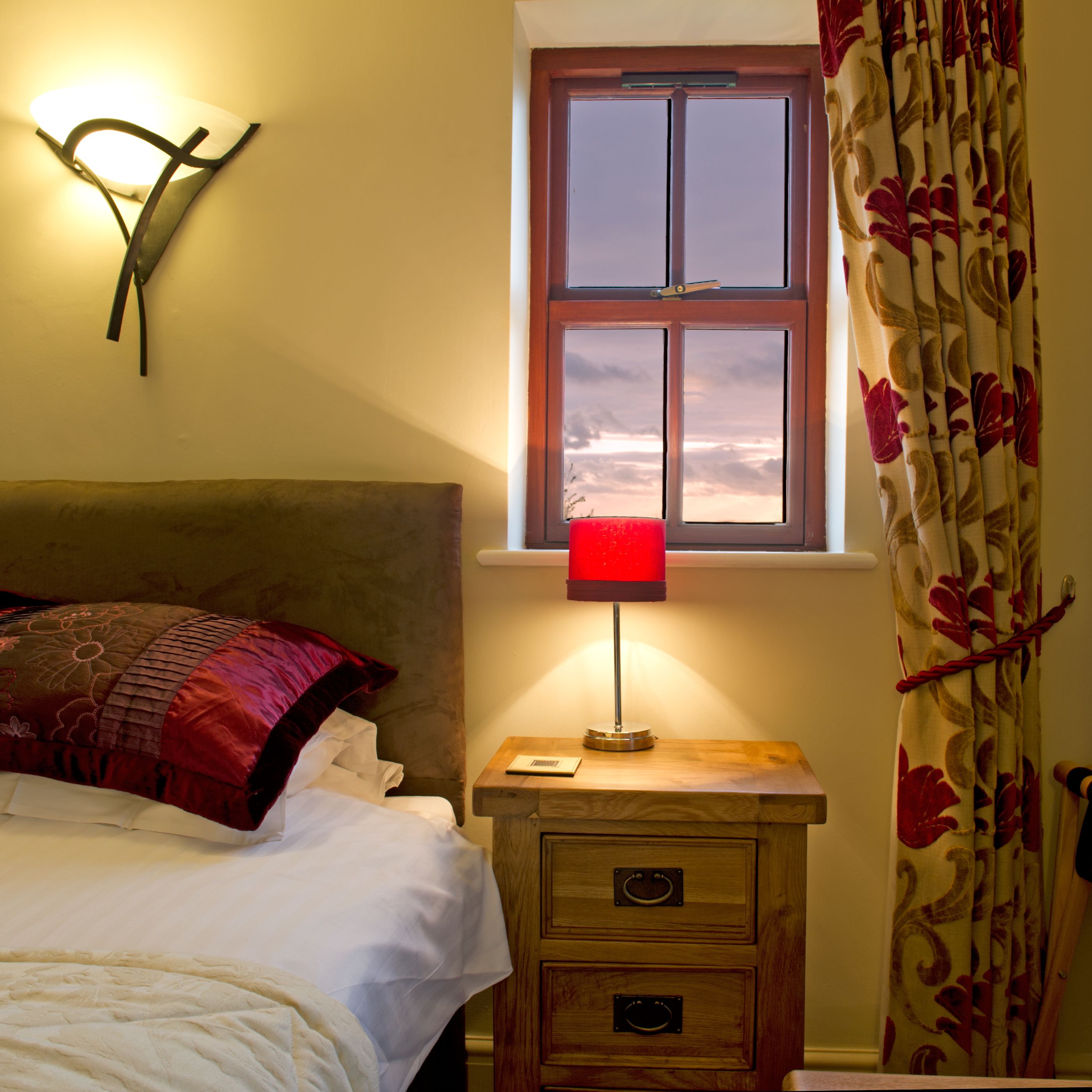 A bedroom with a bed, a nightstand with a lamp, a window with curtains, and a wall-mounted light fixture.