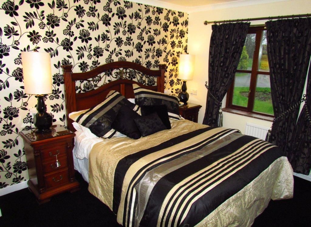 Un dormitorio con papel tapiz floral, cabecera de madera, colcha a rayas, dos lámparas de noche y una ventana con cortinas.