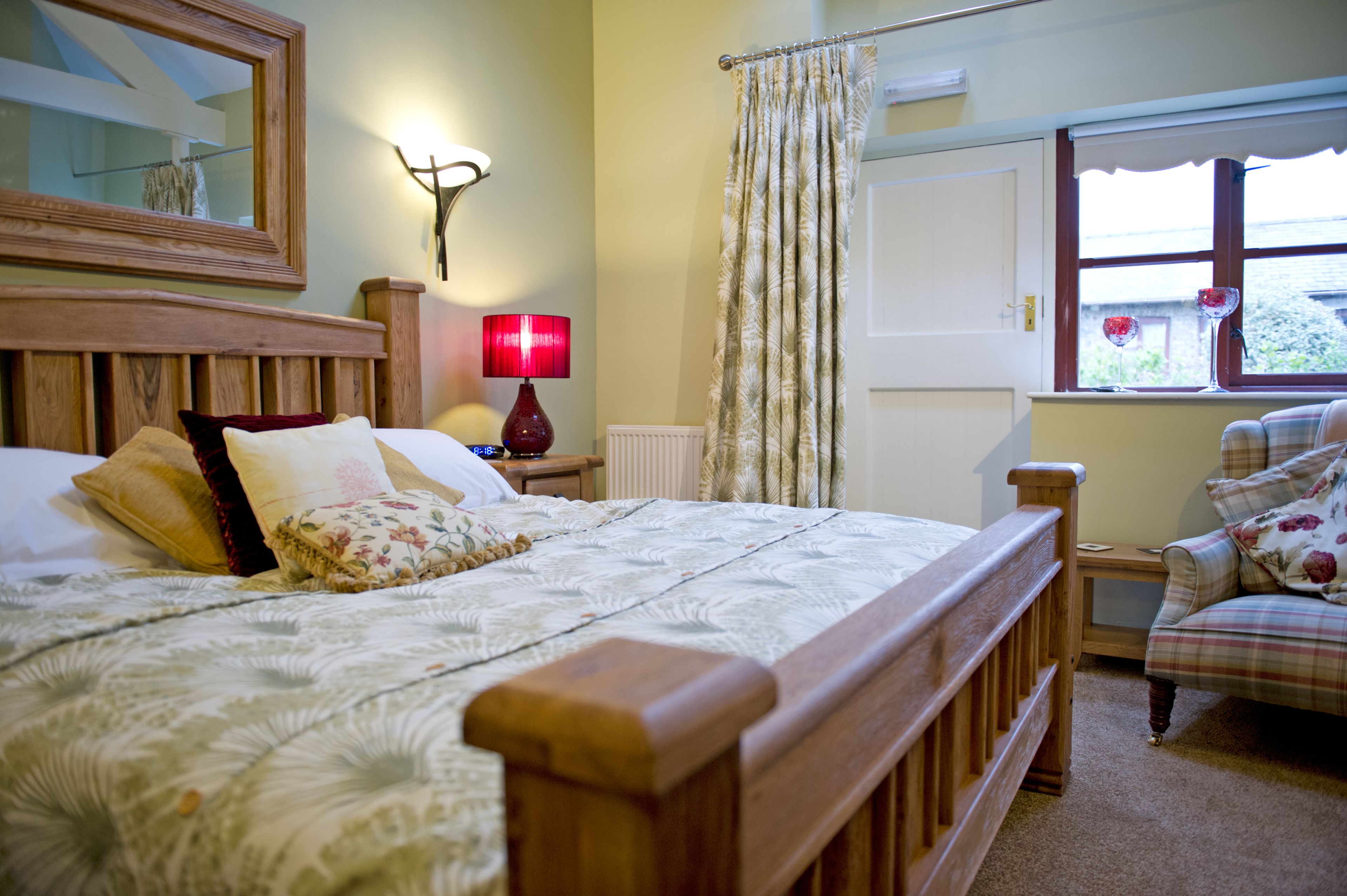 Una camera da letto con un letto in legno, un comodino con una lampada, una sedia, una finestra con tende e uno specchio appeso al muro.