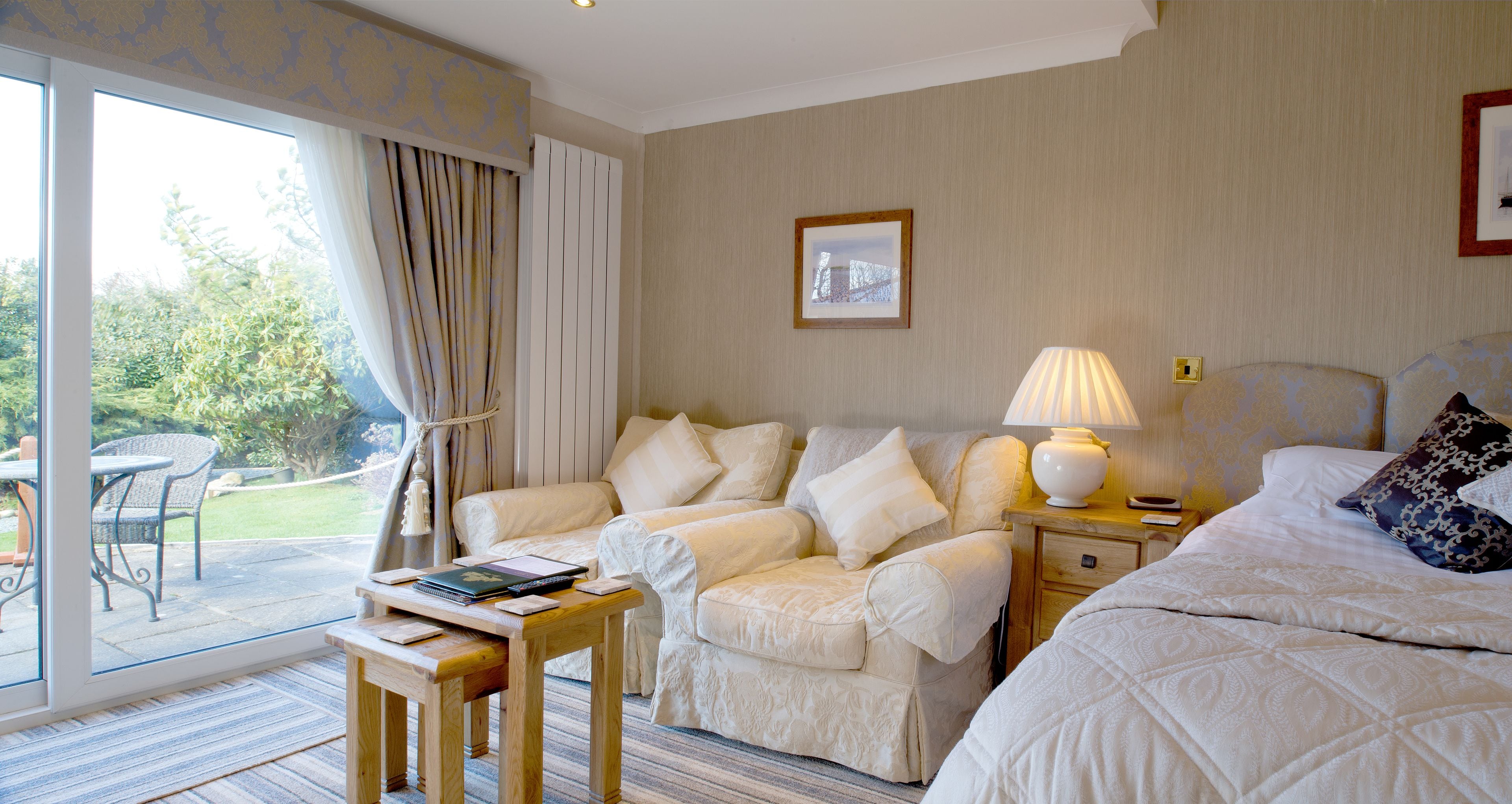 Habitación de hotel con un gran ventanal corredero que da a un patio, una cama con ropa blanca, una mesita de noche con una lámpara y una silla con una computadora portátil.
