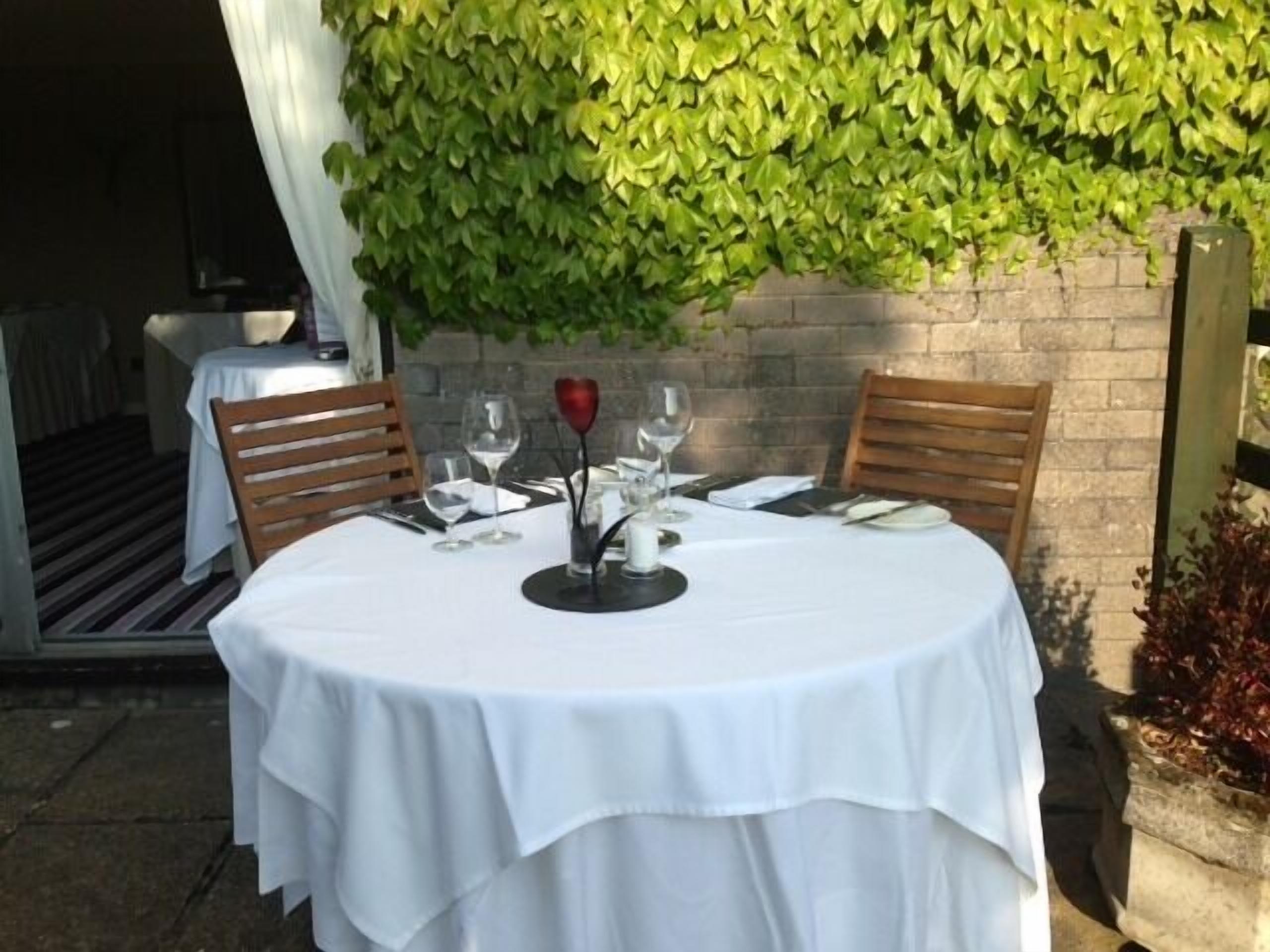 An outdoor dining table with white tablecloths, two wooden chairs, and a vase with a red flower centerpiece.