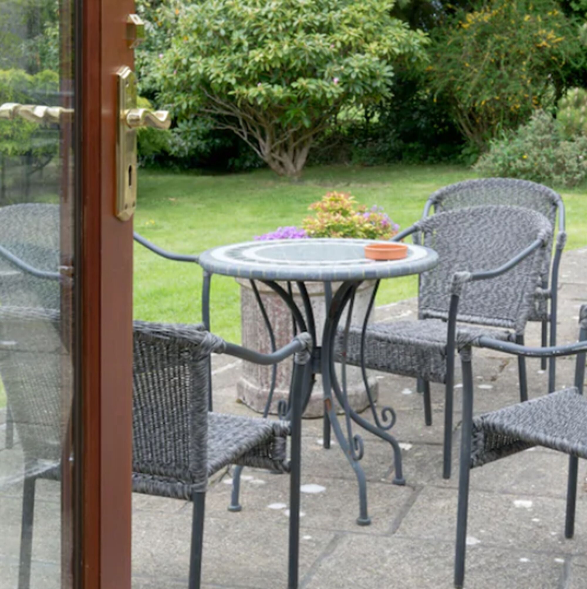 An outdoor patio with wicker chairs and a round table, surrounded by greenery.