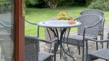 An outdoor patio with wicker chairs and a round table, surrounded by greenery.