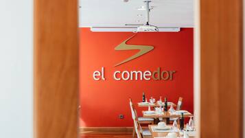 Interior de un restaurante con pared roja y un letrero que dice "el comedor". Mesas dispuestas con vajilla blanca.