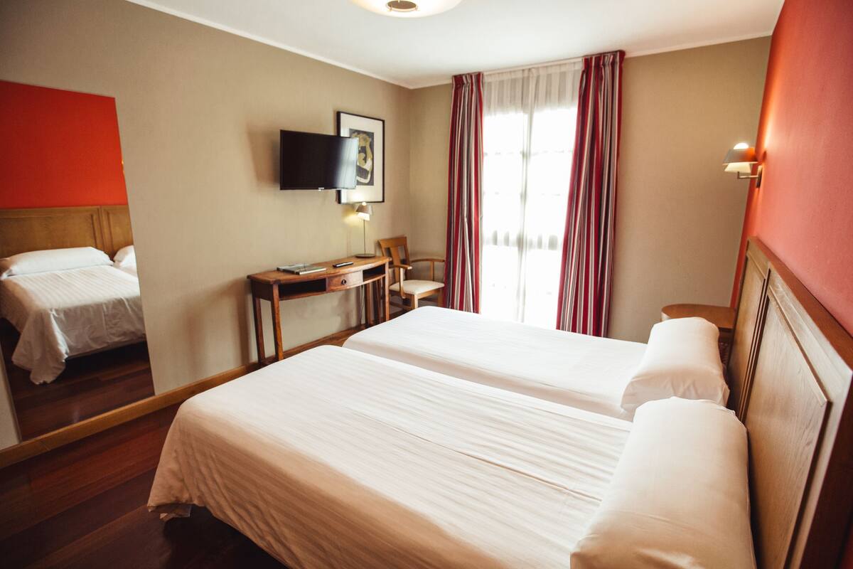 A hotel room with two beds, a desk, a chair, a TV, and a window with curtains.