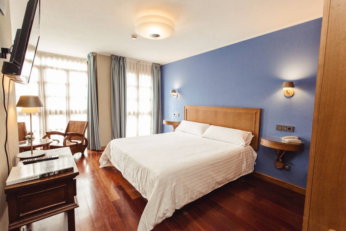 A hotel room with a large bed, wooden headboard, a desk with a lamp, a TV, and a bedside table with a phone.