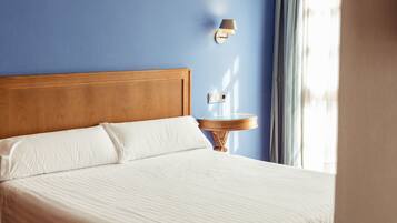 Una cama bien hecha con sábanas blancas, cabecera de madera y una mesita de noche con lámpara.