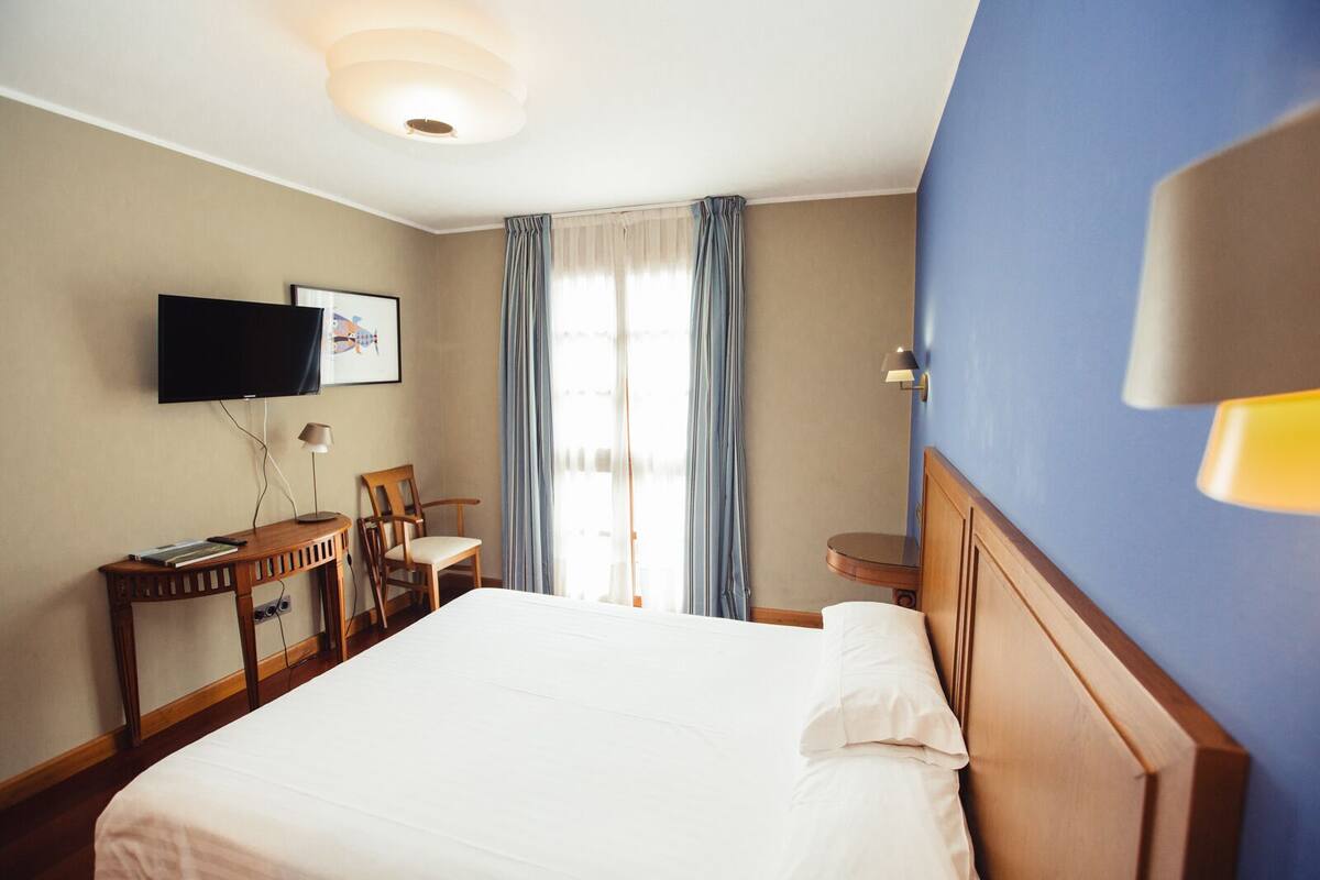 A hotel room with a bed, a desk with a chair, a television, and a window with curtains.