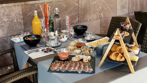 A table set with various food items, including meats, cheeses, and pastries, in a dining area with marble walls and wicker chairs.