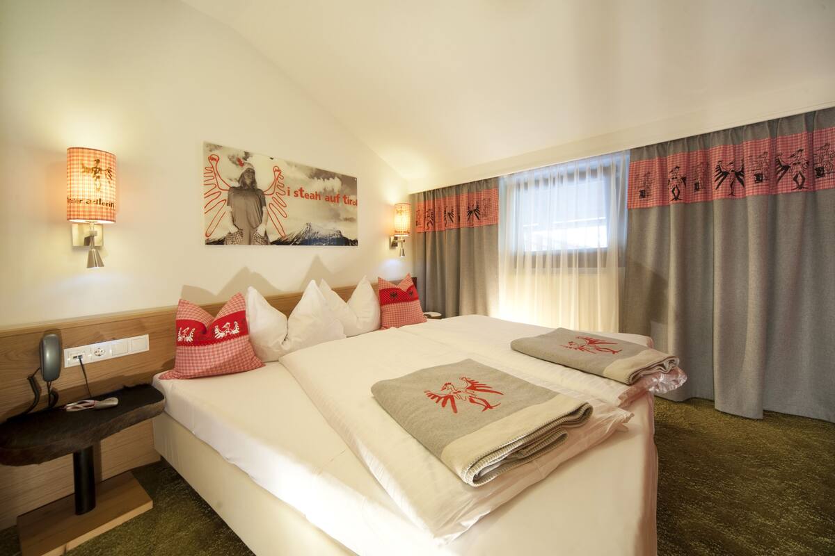 Een hotelkamer met een groot bed, twee nachtkastjes, een bureau en een stoel. Er zijn twee wandlampen, een schilderij en een raam met gordijnen.