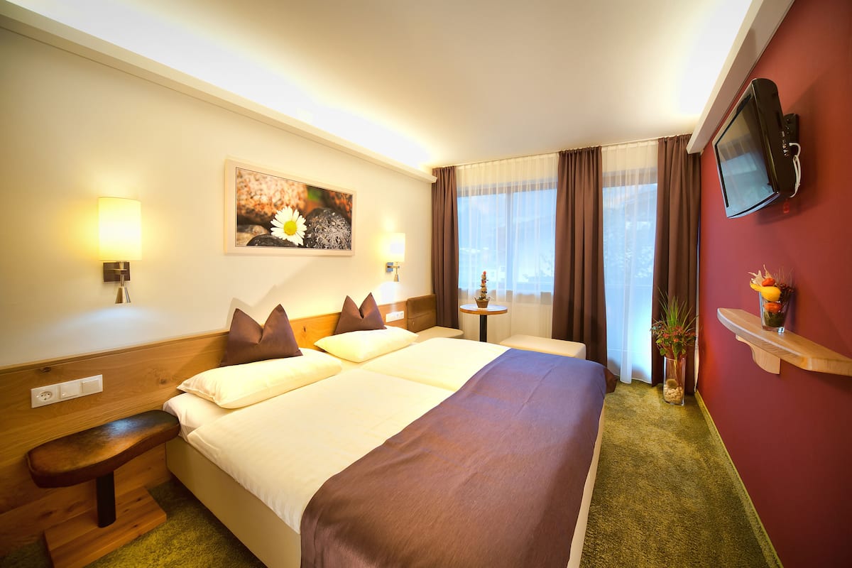 Een hotelkamer met een groot bed, nachtlampjes, een klein tafeltje, een aan de muur bevestigde televisie en een raam met gordijnen.