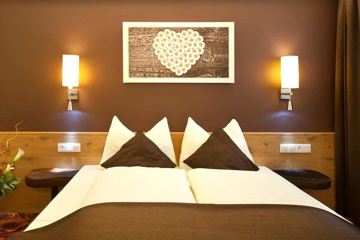Een hotelkamer met een bed, een vaas met bloemen, een hartvormig kunstwerk en twee wandlampen.