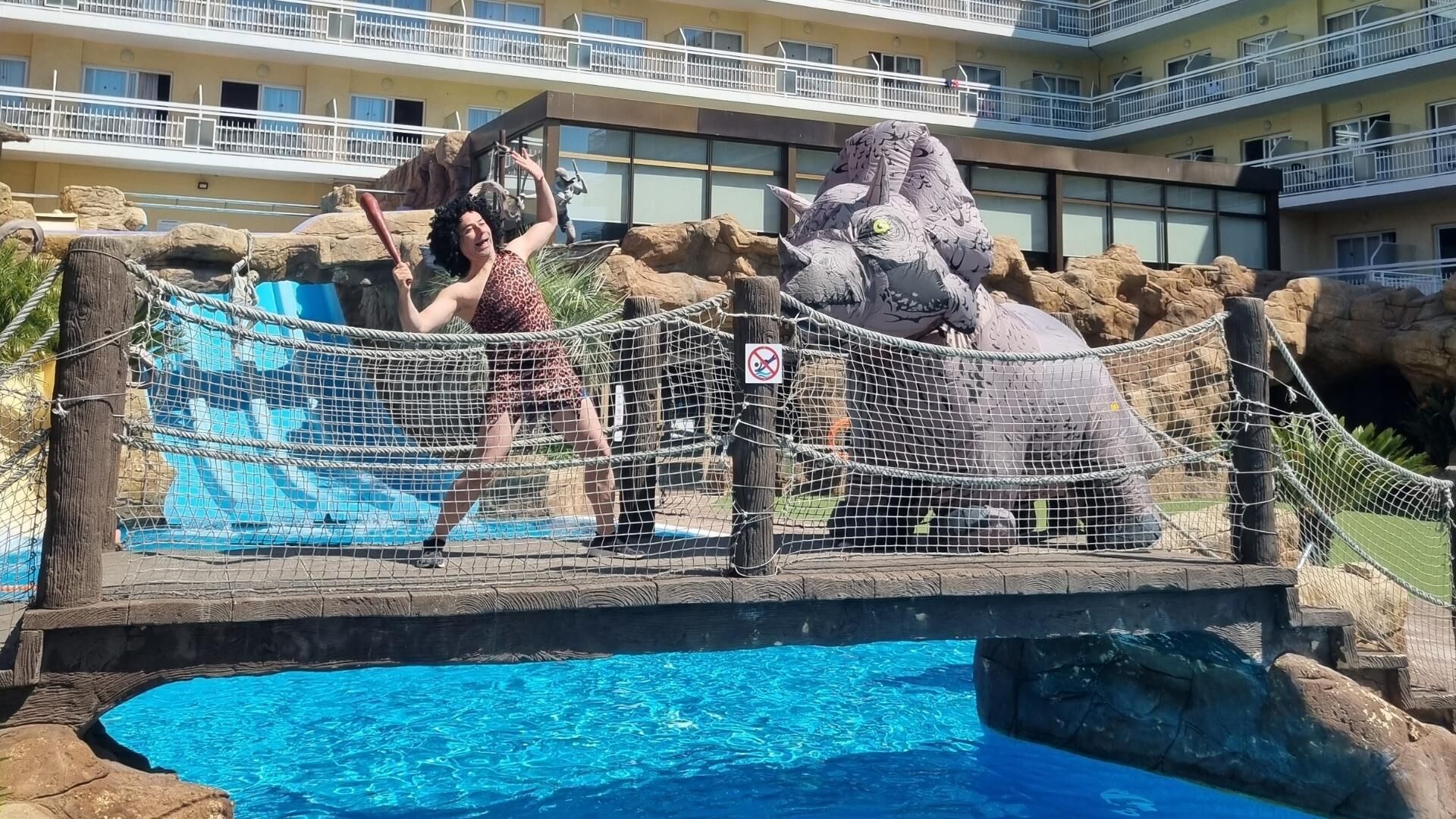 A pool area with a rope bridge, a large rock formation, and a building with balconies in the background.