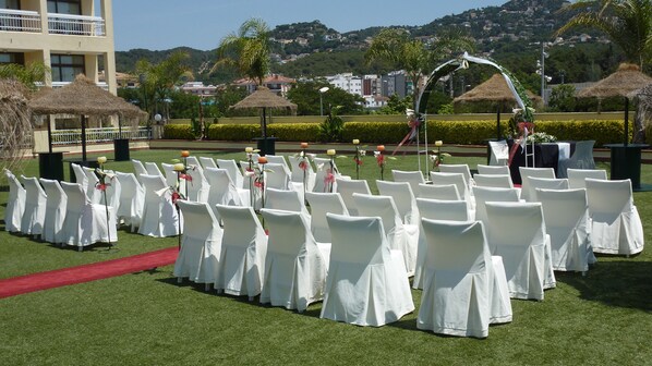Rows of white chairs with red carpet, a stage with a white arch, and a backdrop of a residential area with hills.
