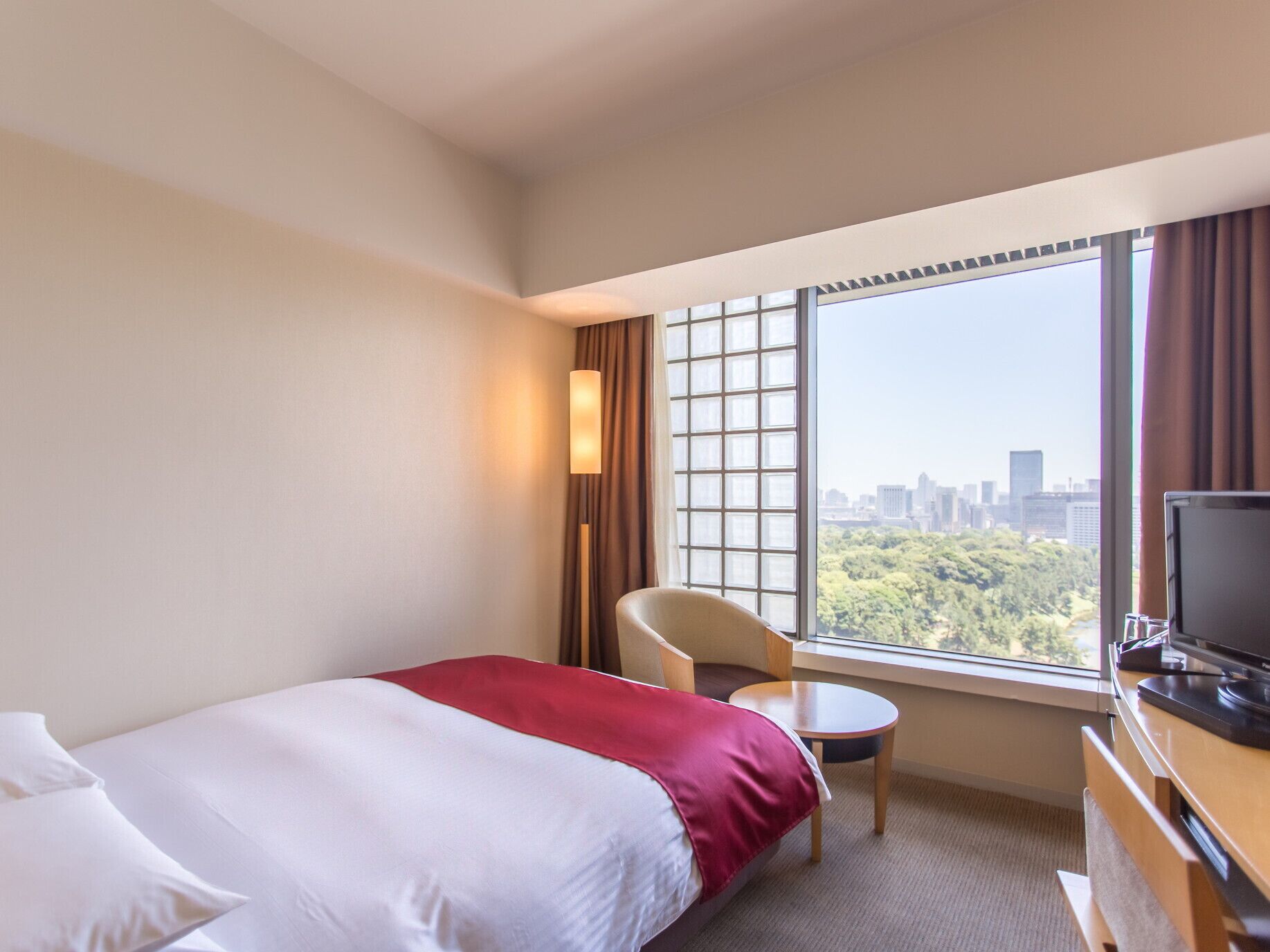 A hotel room with a bed, a chair, a small table, a TV, and a window with a city view.