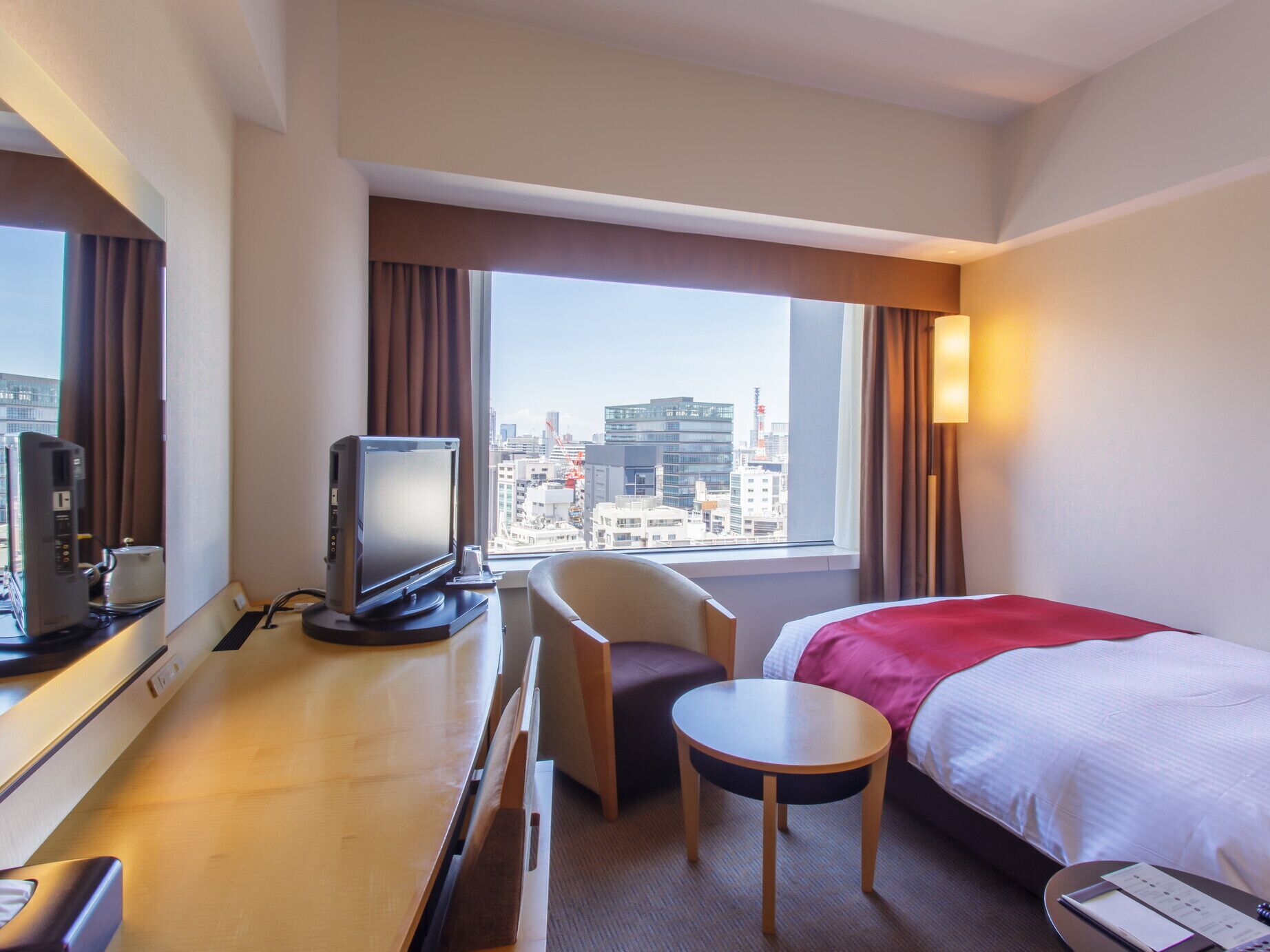 A hotel room with a bed, a desk with a television, a chair, a small table, and a view of the cityscape.