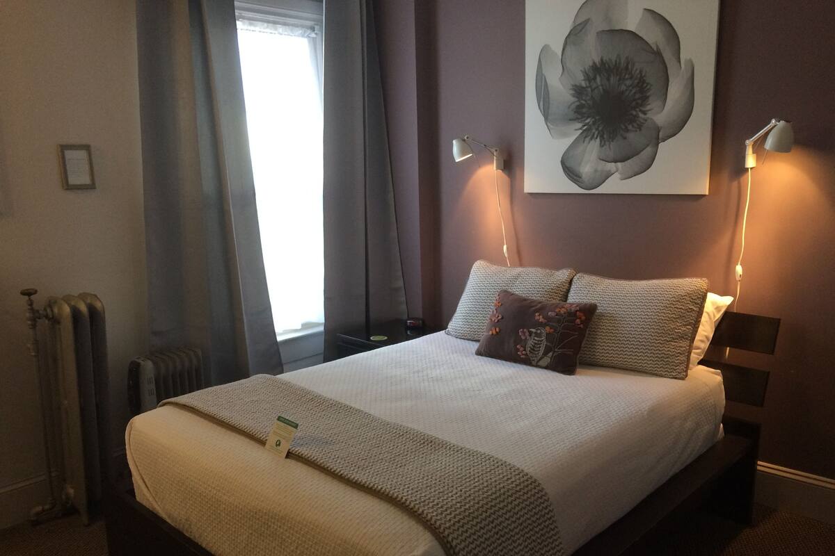 Un lit bien fait, avec une couette beige et des oreillers, deux lampes de chevet, un radiateur et une grande œuvre d’art florale accrochée au mur.