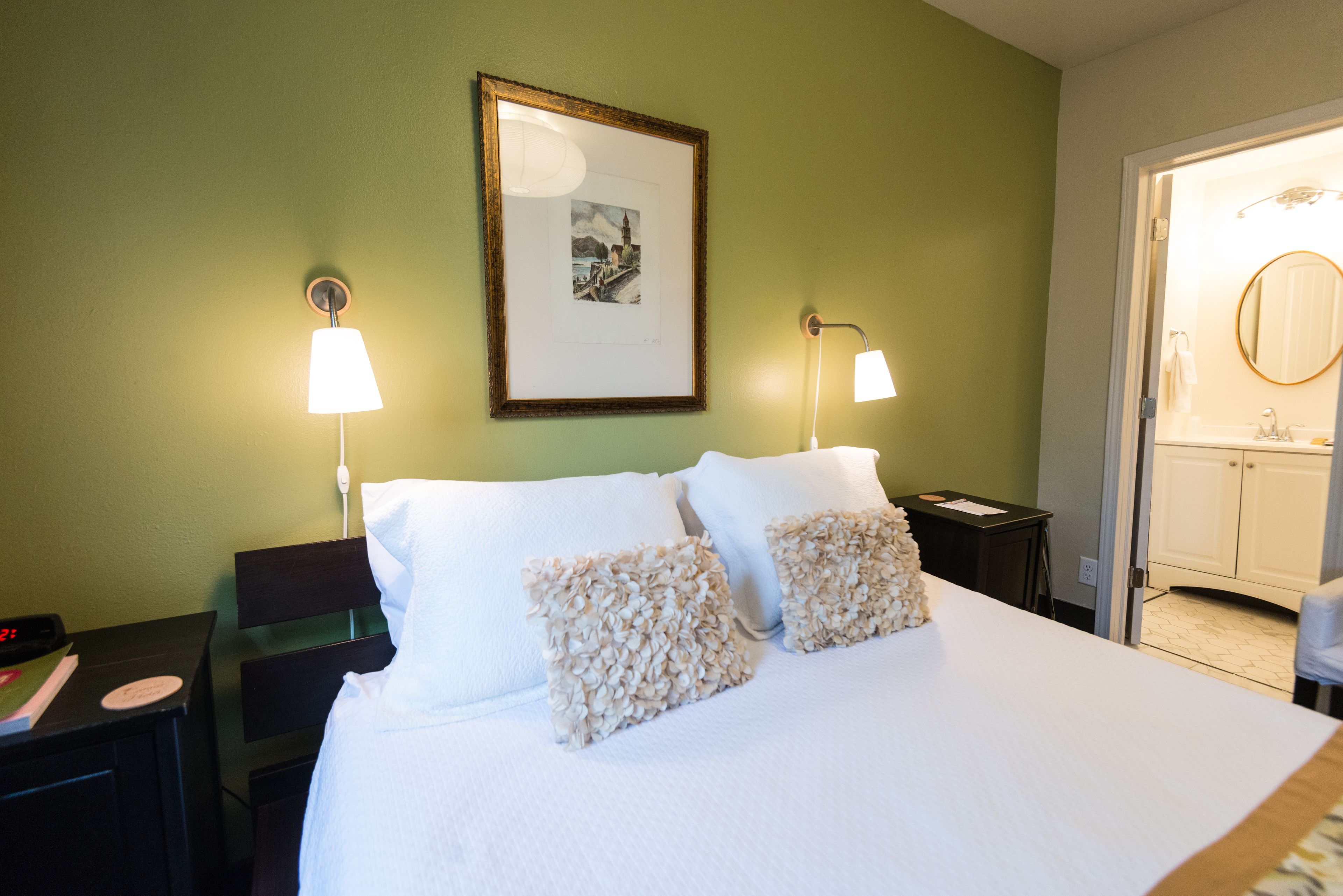 Una cama bien hecha con sábanas blancas y almohadas decorativas, un cuadro en la pared y una mesita de noche con una lámpara.