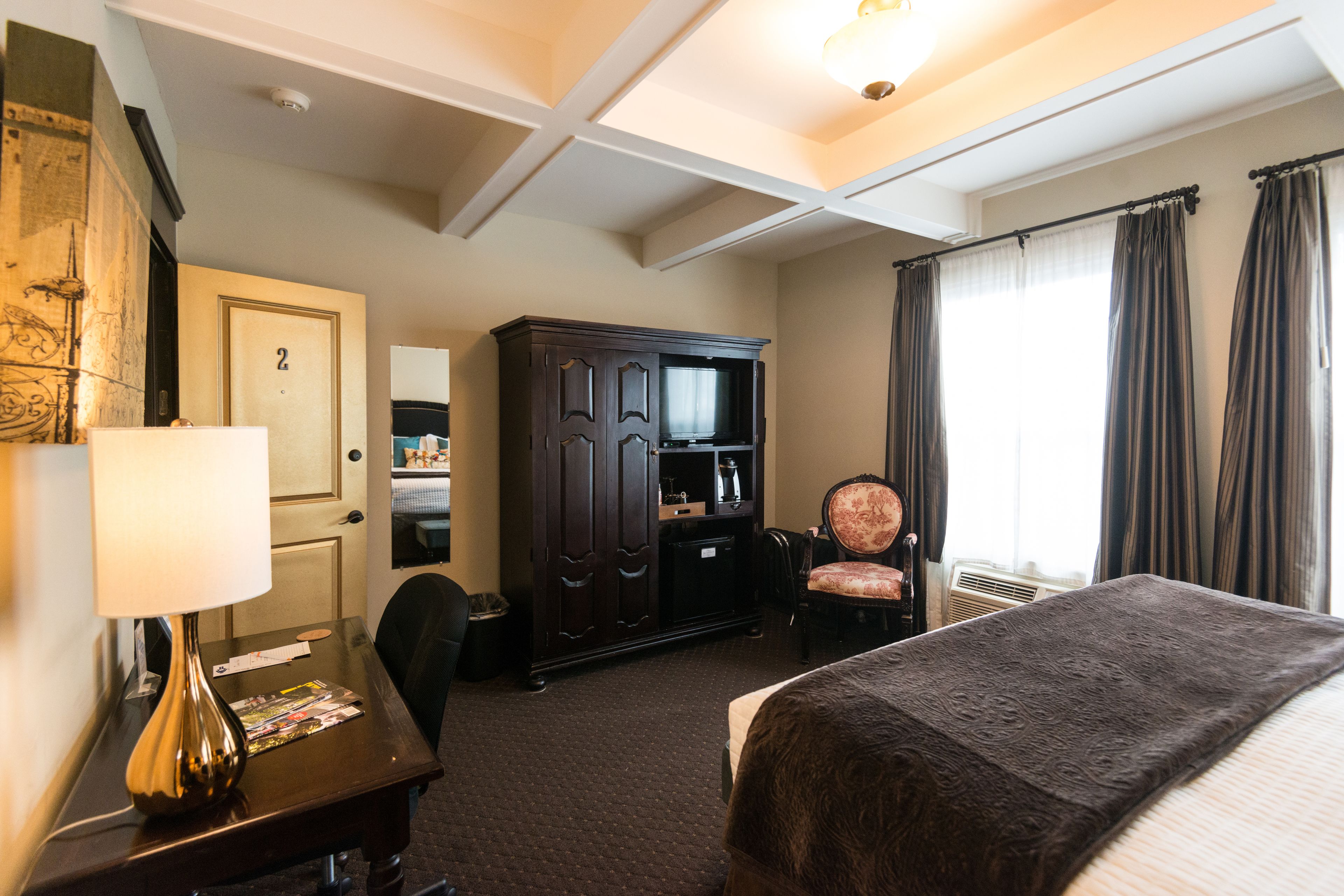 A hotel room with a bed, a desk with a lamp, a wardrobe, a television, a chair, and a window with curtains.