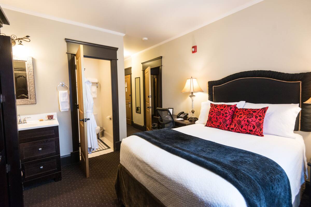 Une chambre d’hôtel comprenant un lit, une table de chevet, une lampe, un miroir, une salle de bain avec un lavabo et des toilettes, et une porte donnant sur une autre pièce.