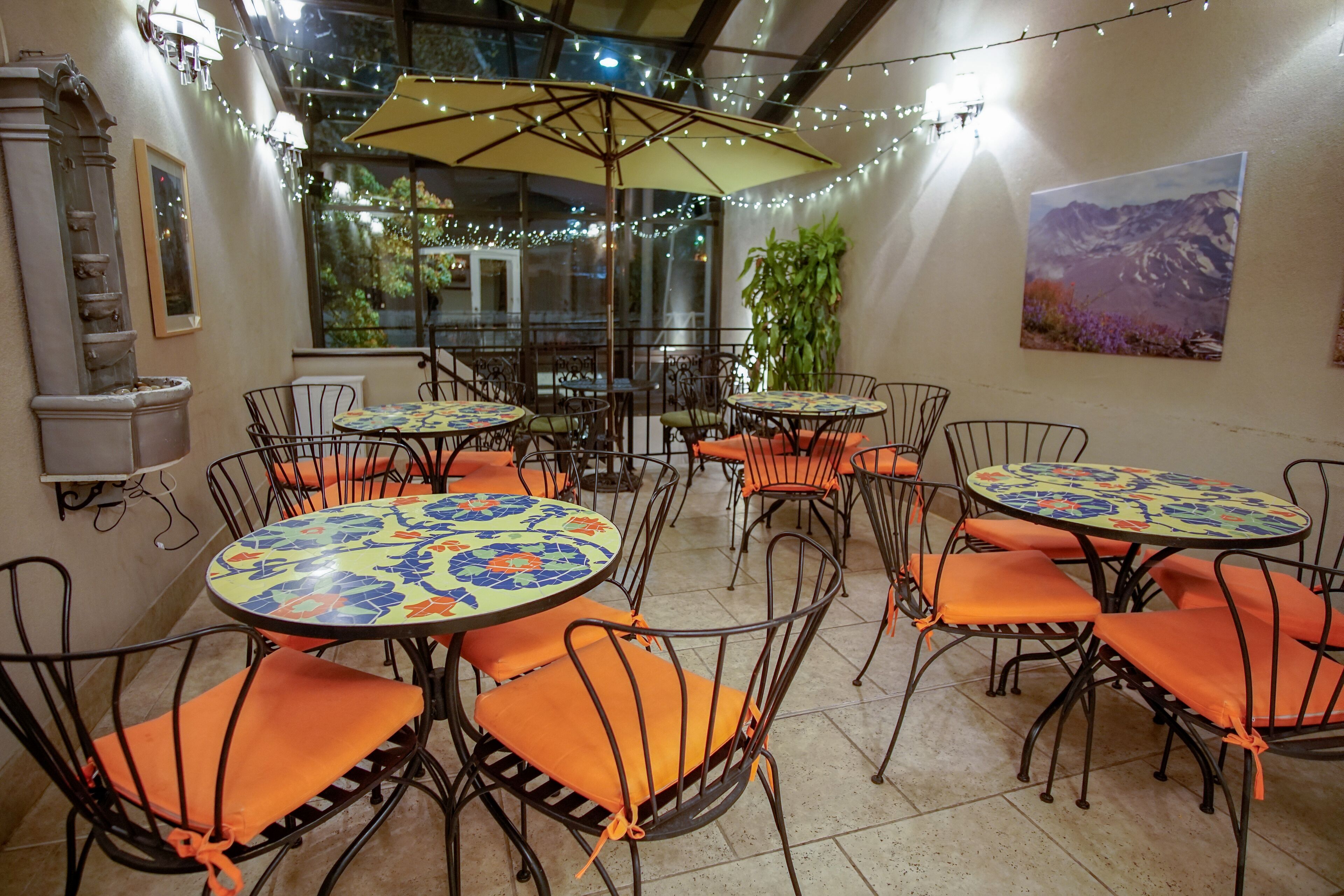 An outdoor patio area with round tables and chairs, string lights, and a mountain landscape painting on the wall.