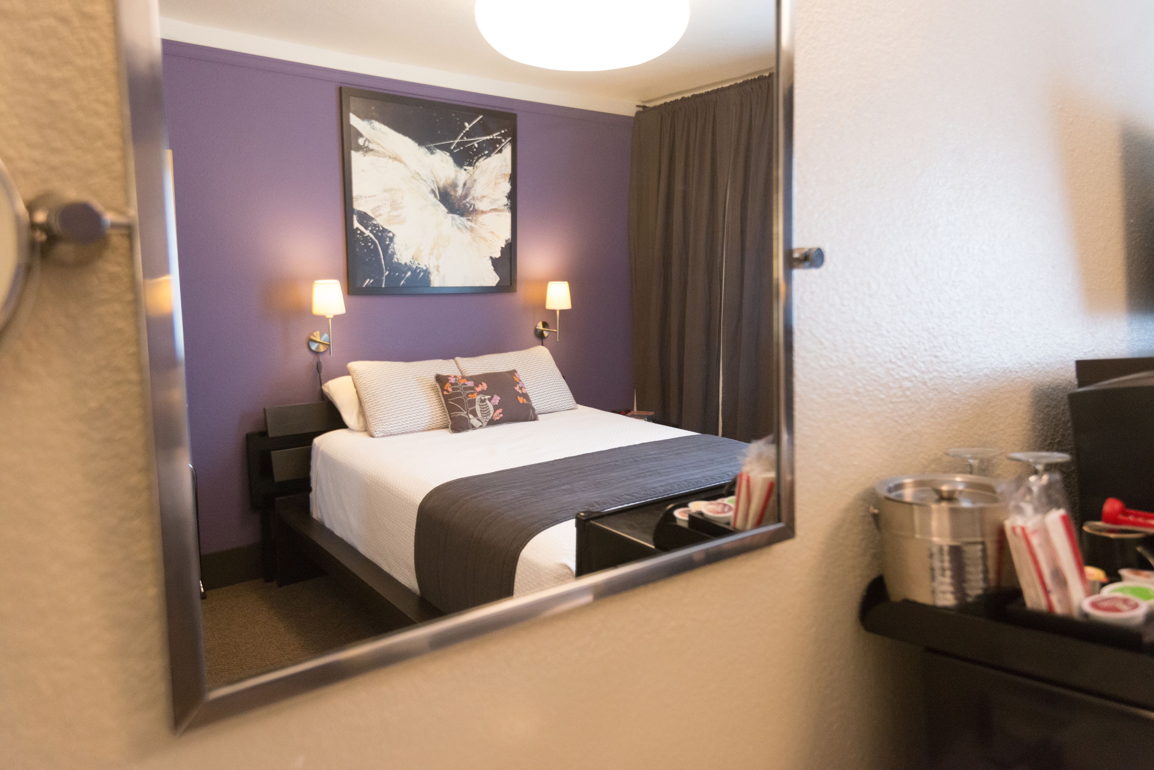 Una camera d'albergo con un letto, un comodino, una lampada, un quadro appeso alla parete e uno specchio che riflette la stanza.