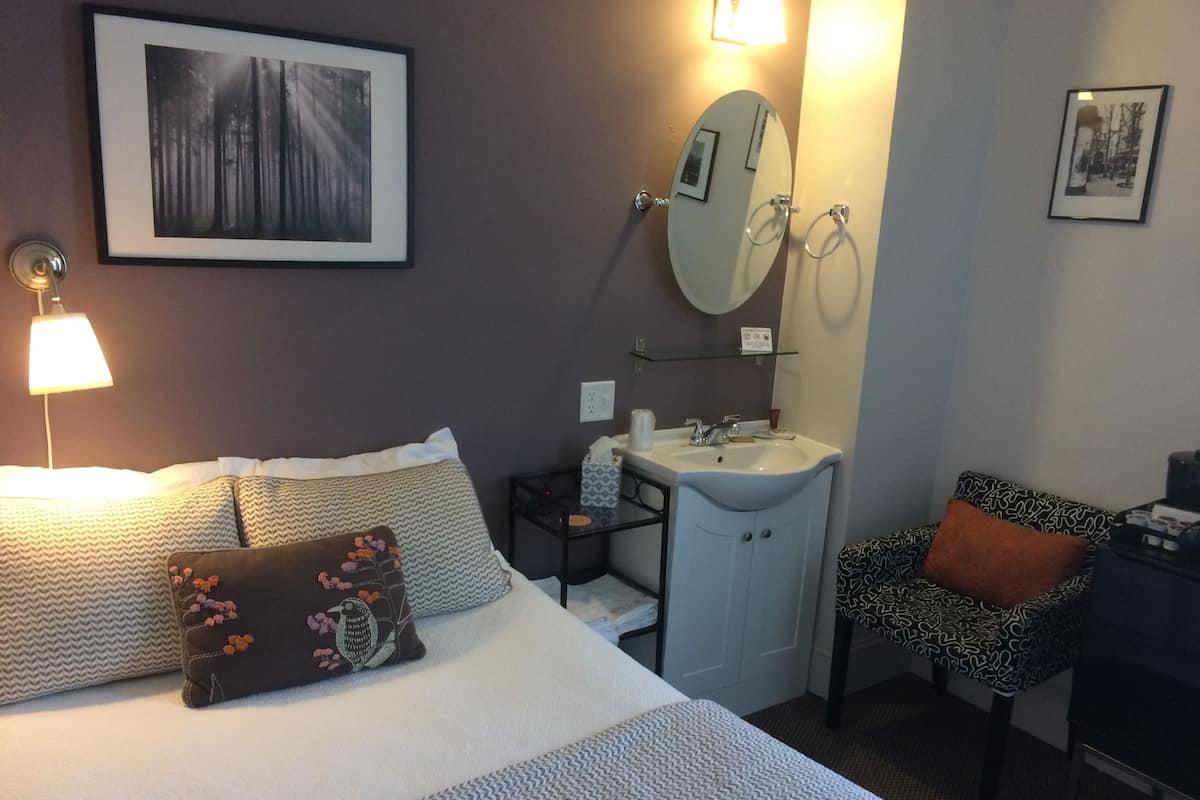 Une chambre d’hôtel comprenant un lit, une table de chevet, un petit bureau, une chaise, une salle de bain avec un lavabo et un miroir, et un tableau encadré au mur.
