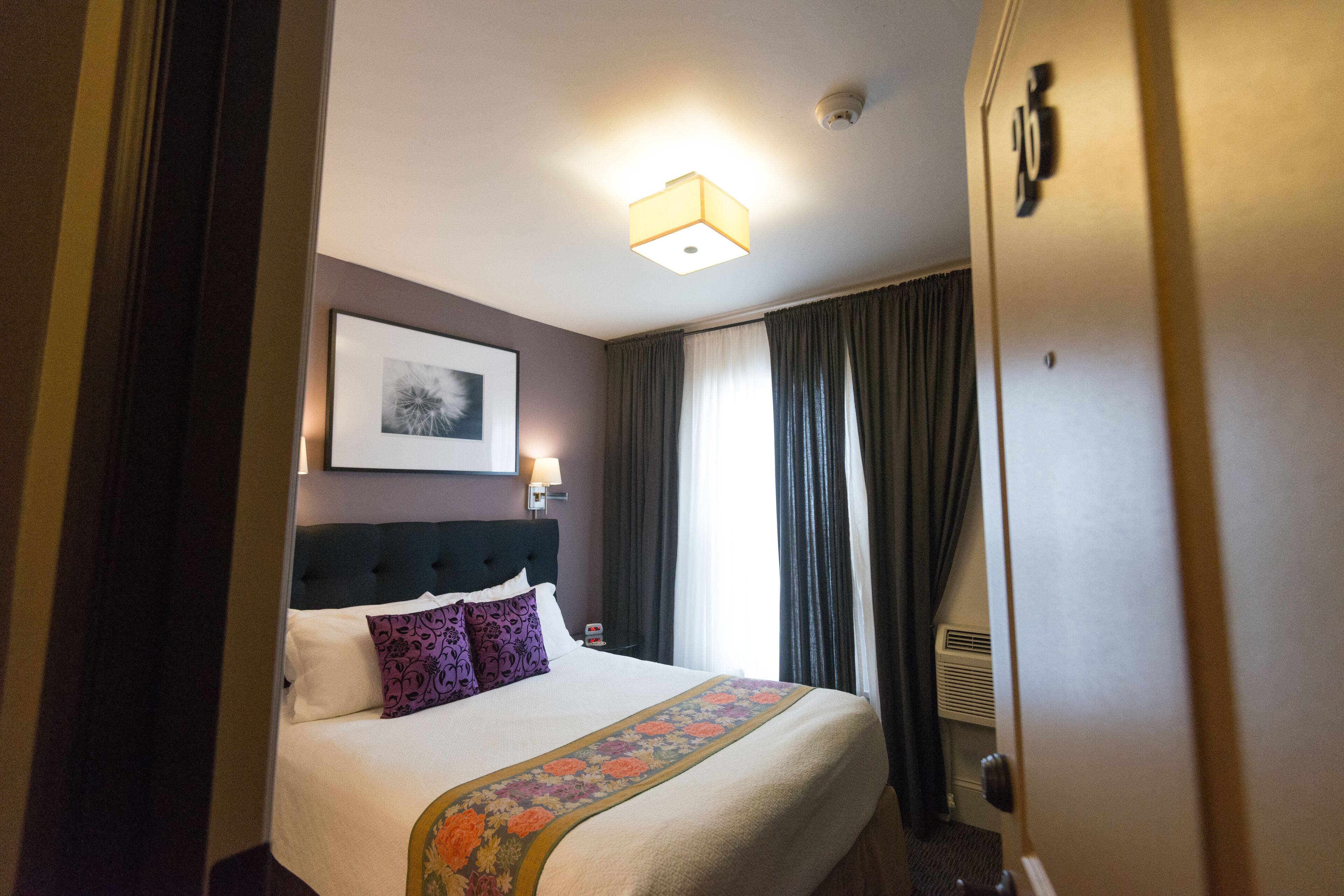 A hotel room with a neatly made bed, a headboard, a framed picture on the wall, and a window with curtains.