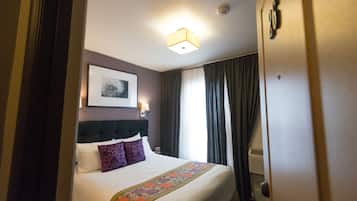 A hotel room with a neatly made bed, a headboard, a framed picture on the wall, and a window with curtains.