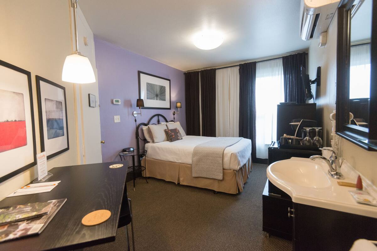 Une chambre d’hôtel avec un lit, un bureau, une salle de bain avec un lavabo et un miroir, et des tableaux encadrés accrochés au mur.
