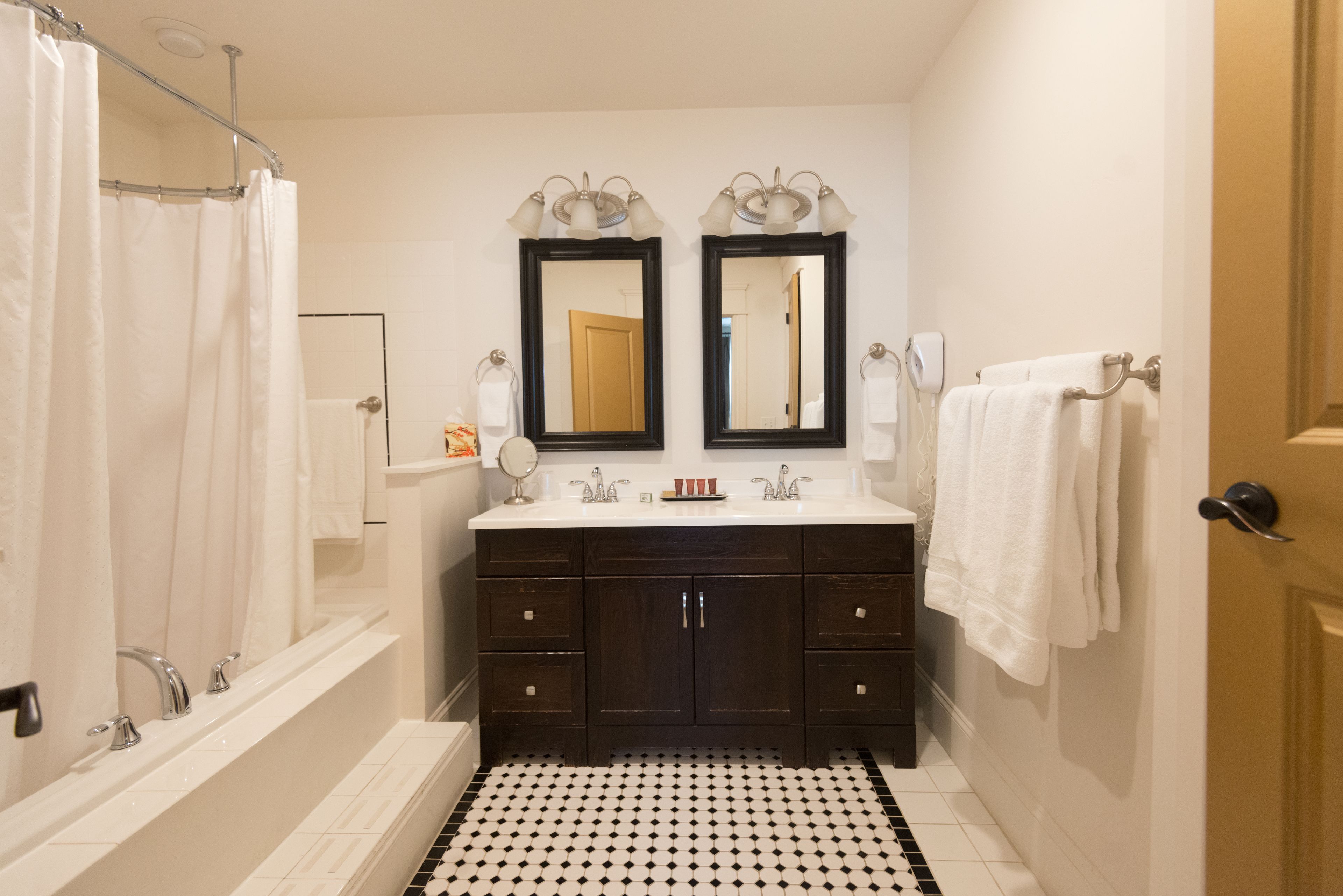 A bathroom with a bathtub, double sink vanity, two mirrors, and a black and white tiled floor.