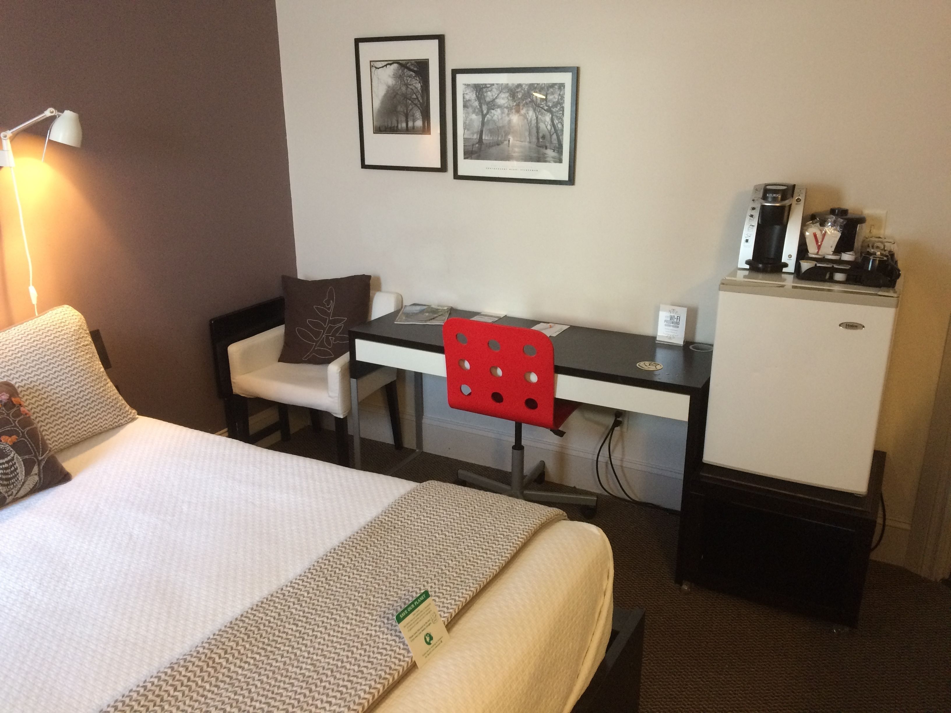 Une chambre d’hôtel comprenant un lit, un bureau, une chaise, un petit réfrigérateur et deux tableaux encadrés accrochés au mur.
