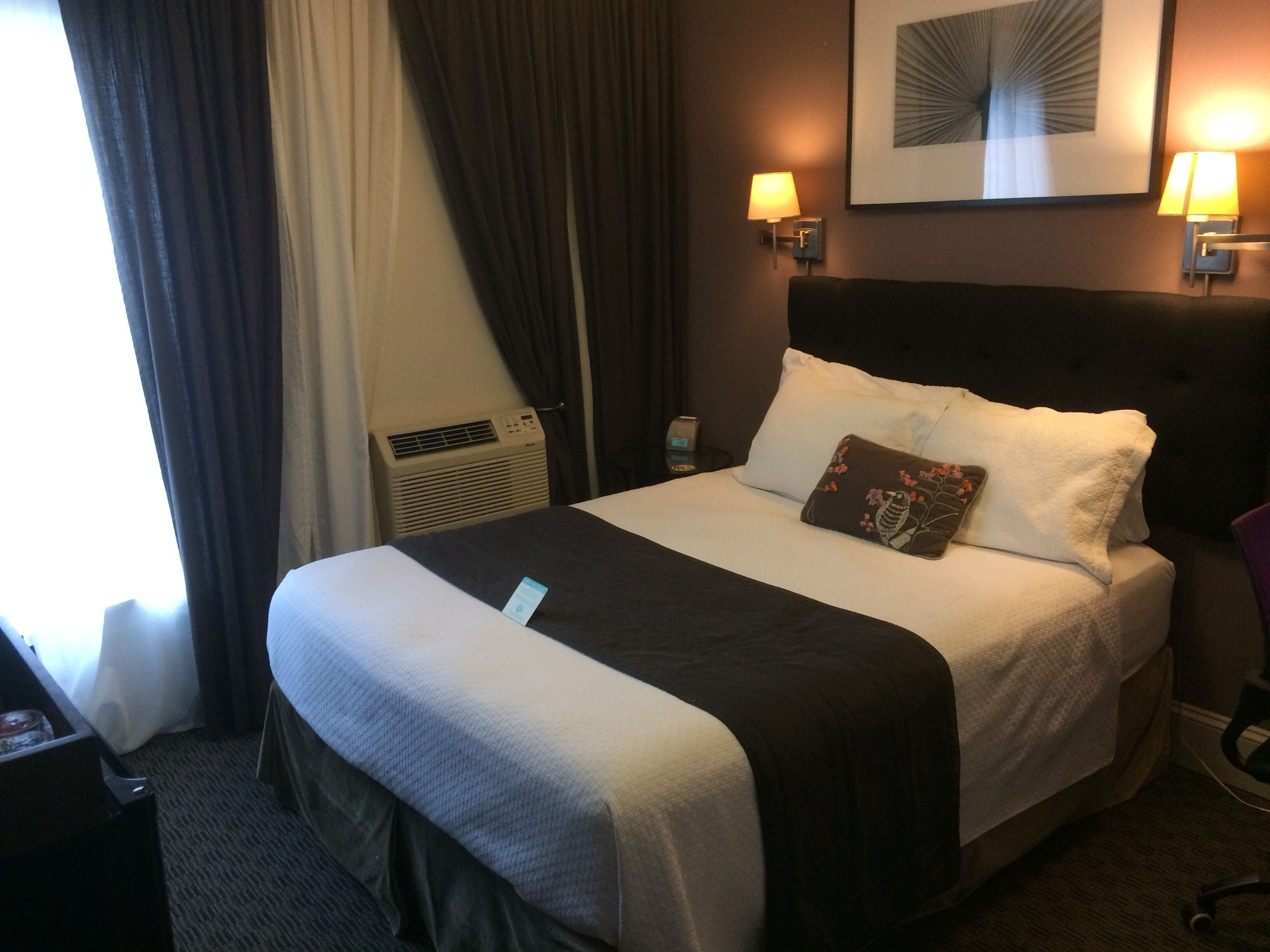 A hotel room with a bed, a nightstand, a lamp, a wall-mounted air conditioner, and a framed picture on the wall.