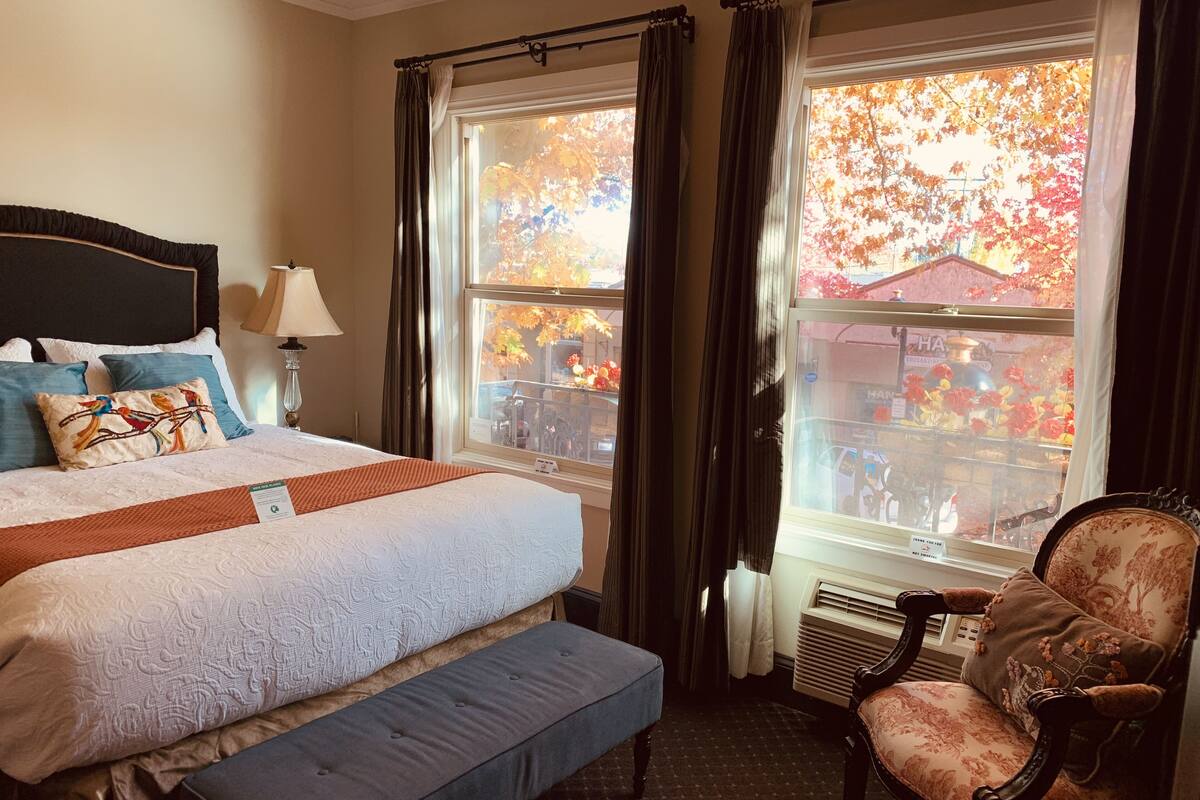 Une chambre d’hôtel avec un grand lit, une banquette, une chaise et une fenêtre donnant sur le paysage automnal.