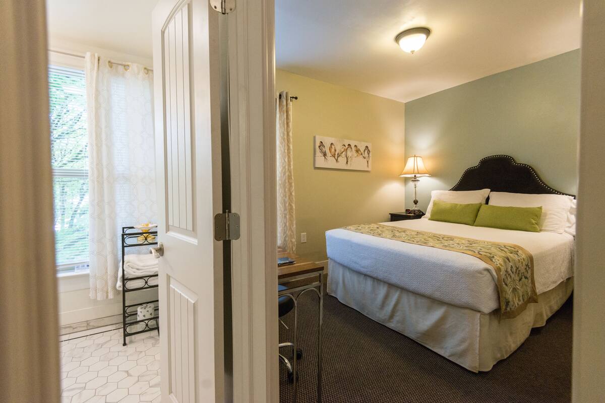 Une chambre d’hôtel avec un lit, une table de chevet, une lampe, une fenêtre avec des rideaux et un mur orné d’un tableau représentant un oiseau.
