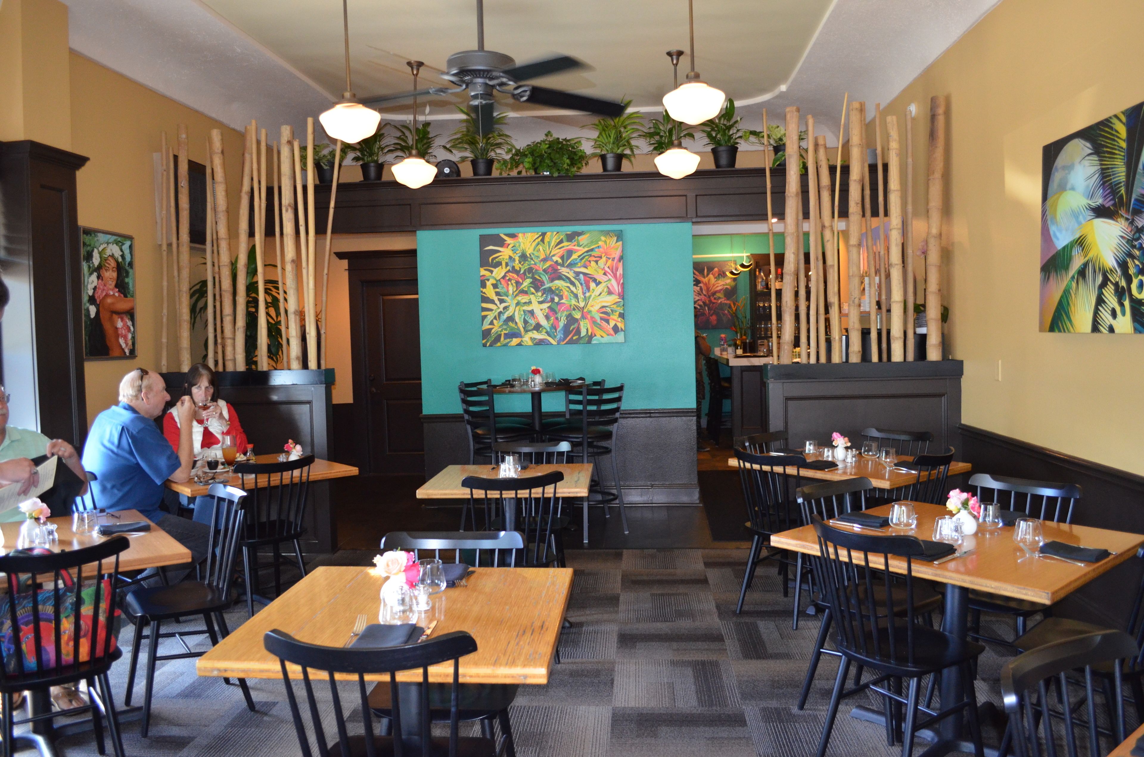 A restaurant with wooden tables and black chairs, a ceiling fan, and potted plants on the walls.