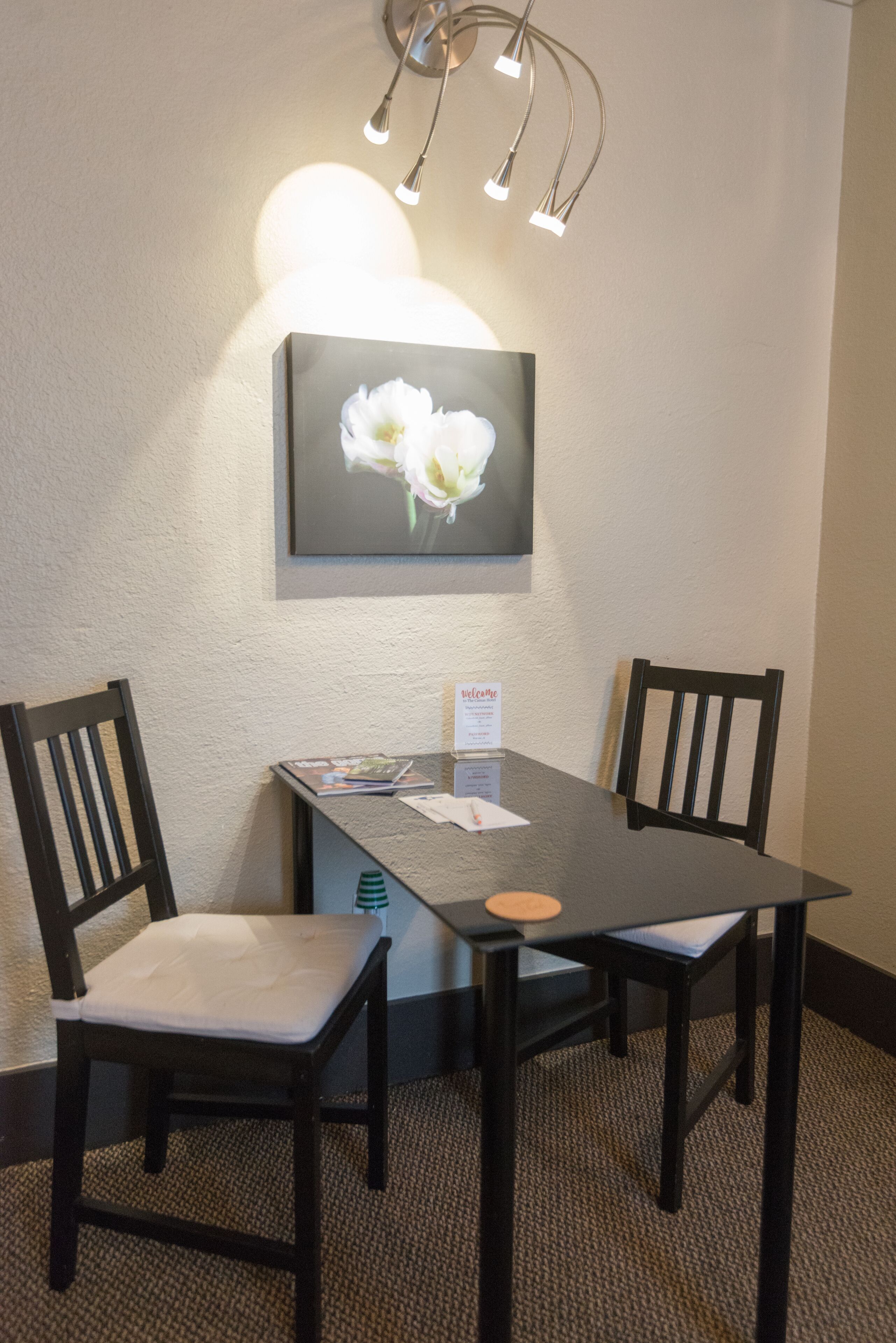 A hotel room with a dark wooden table, two chairs, a framed floral artwork on the wall, and a small welcome sign.