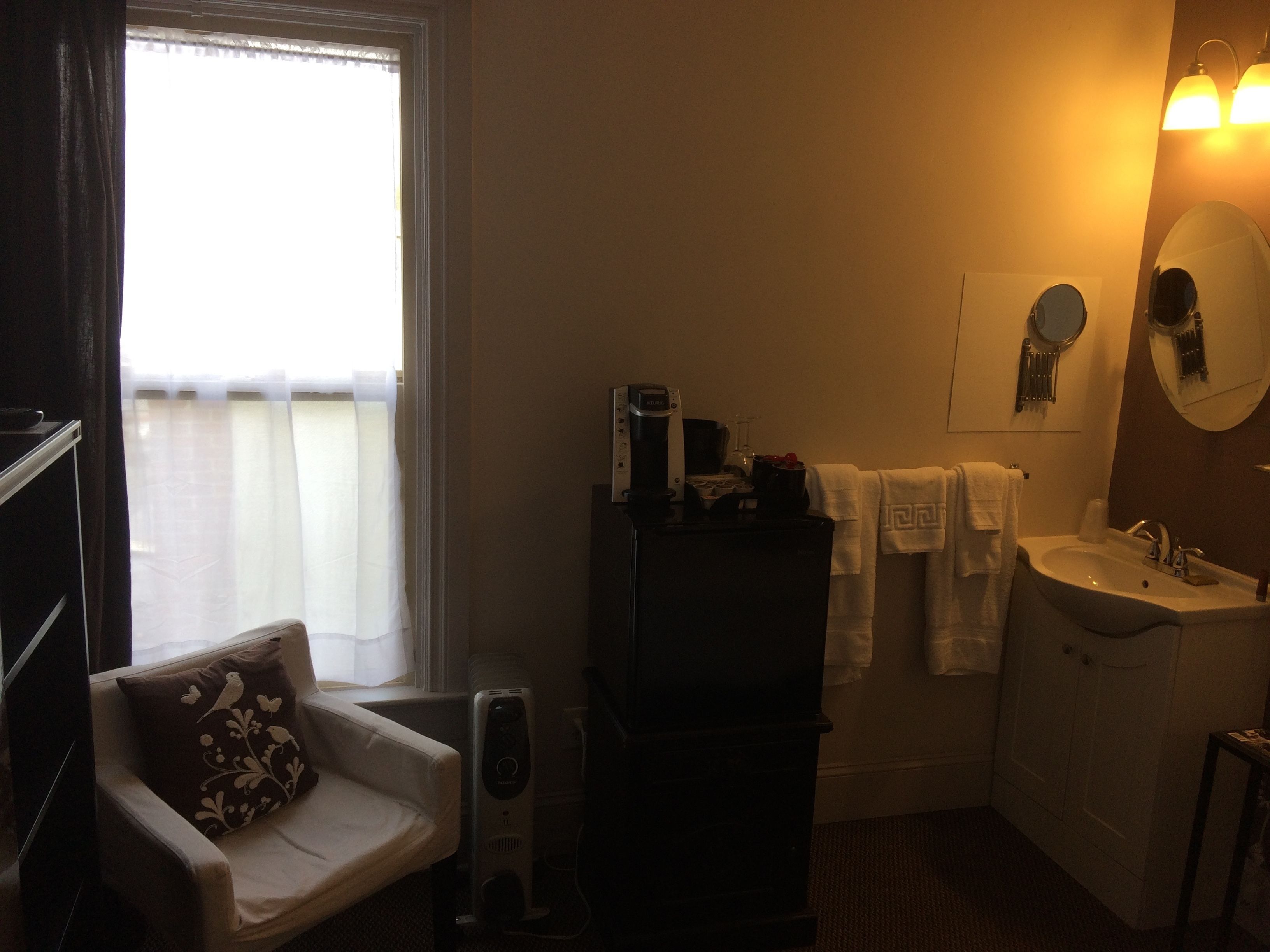 Ein Hotelzimmer mit einem Bett, einem Sessel mit Zierkissen, einem Nachttisch mit Lampe, einem Schminktisch, einem Spiegel, einem Waschbecken und einem Fenster mit Vorhängen.