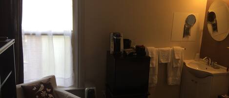 Habitación de hotel con cama, sillón con almohada decorativa, mesita de noche con lámpara, cómoda, espejo, lavamanos y ventana con cortinas.