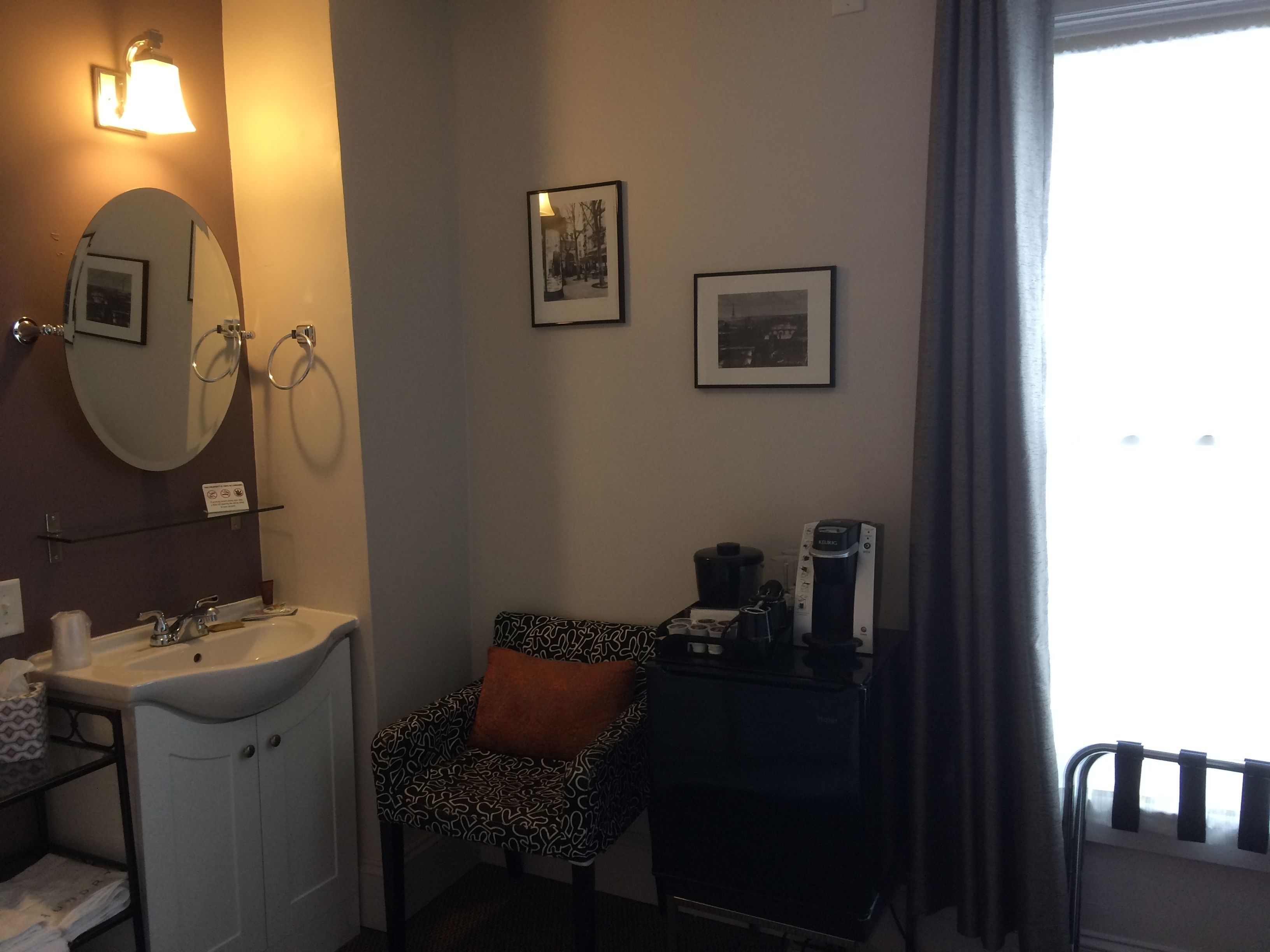 A hotel room with a sink, mirror, towel rack, two framed pictures, a chair with a patterned cushion, a coffee maker, and a window with curtains.