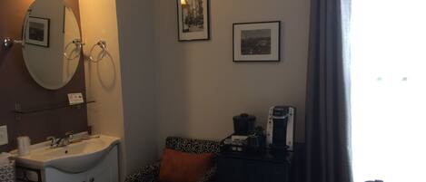 A hotel room with a sink, mirror, towel rack, two framed pictures, a chair with a patterned cushion, a coffee maker, and a window with curtains.