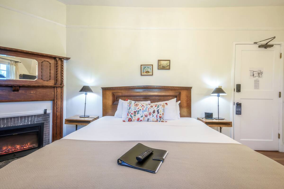 A hotel room with a large bed, a fireplace, two bedside lamps, and a door with a sign.