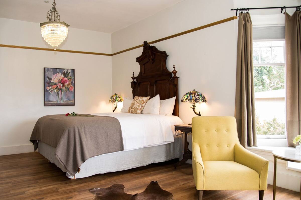 A bedroom with a large bed, a wooden headboard, a chandelier, a painting, and a window with curtains.