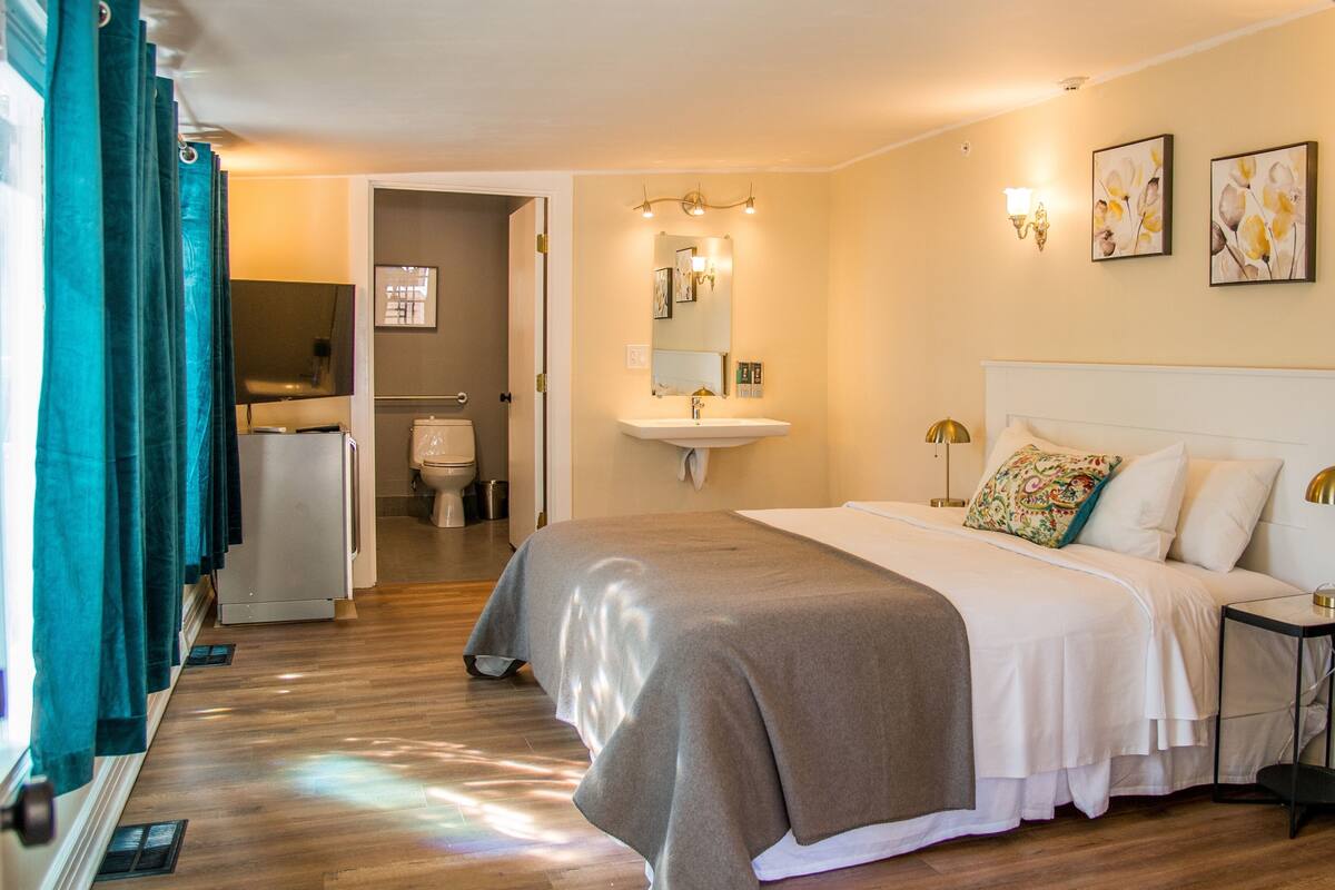 A hotel room with a bed, bedside table, lamp, and a bathroom visible through an open door.