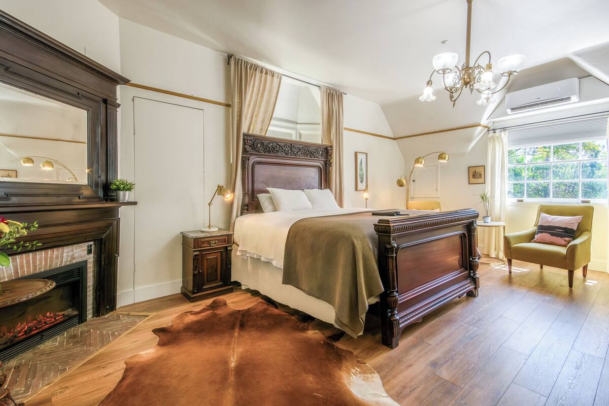 A bedroom with a large bed, a fireplace, a chandelier, and a view of greenery outside.