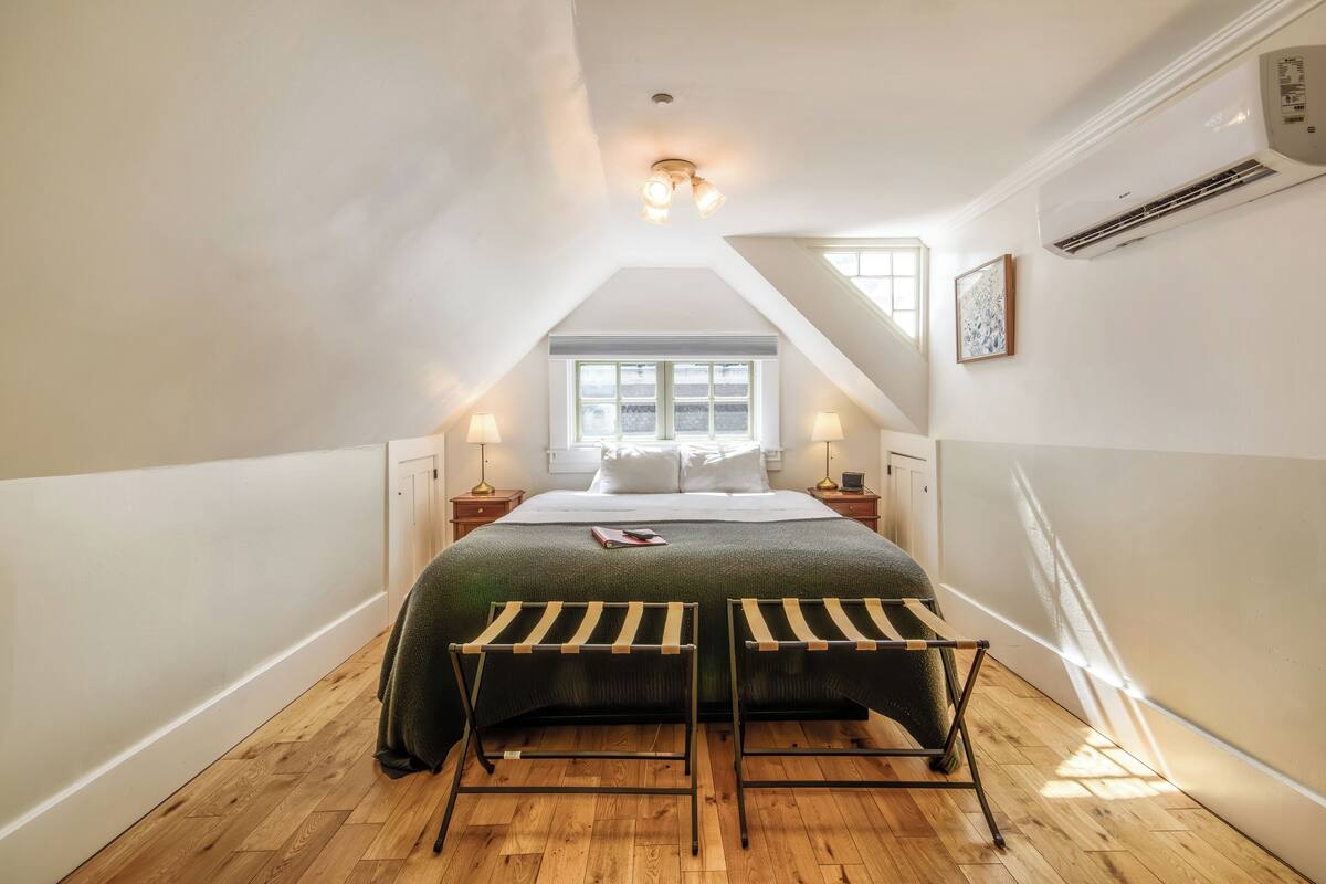 A bedroom with a sloped ceiling, a bed with a green blanket, two bedside tables with lamps, and a window with blinds.