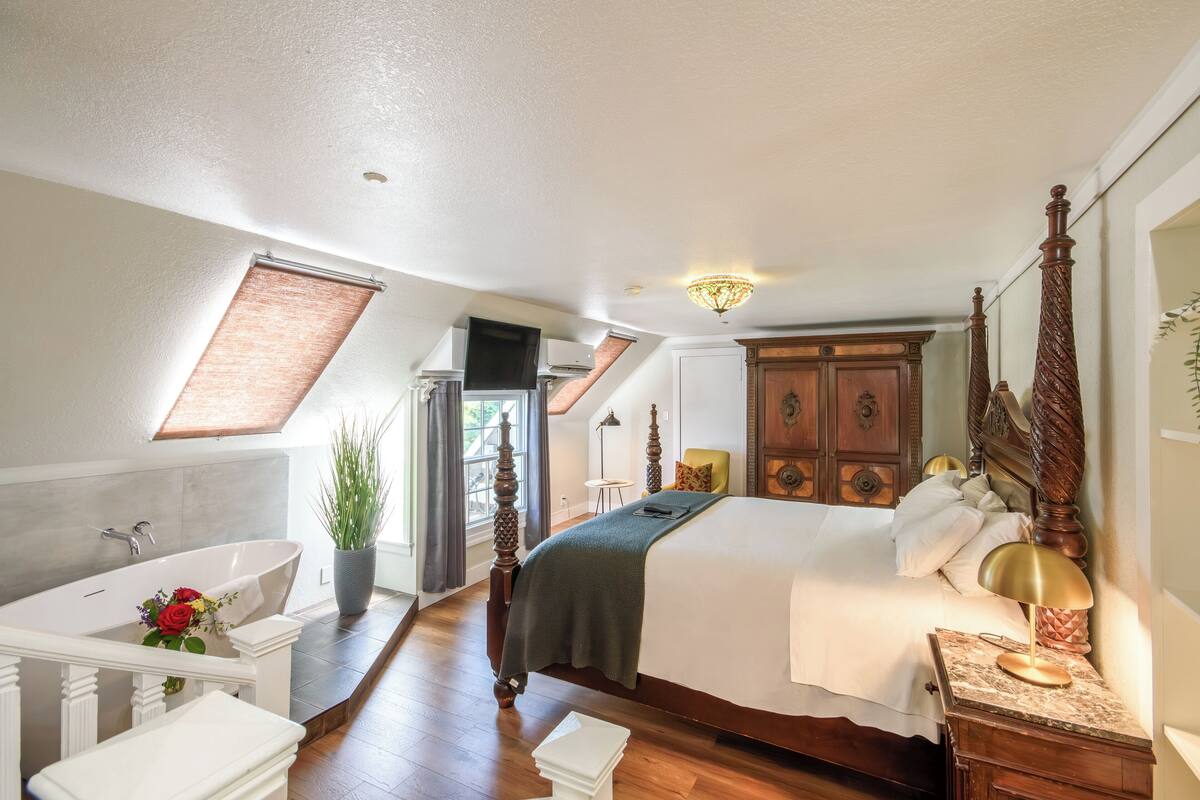 A bedroom with a large bed, a wooden wardrobe, a bathroom with a bathtub, and a skylight.